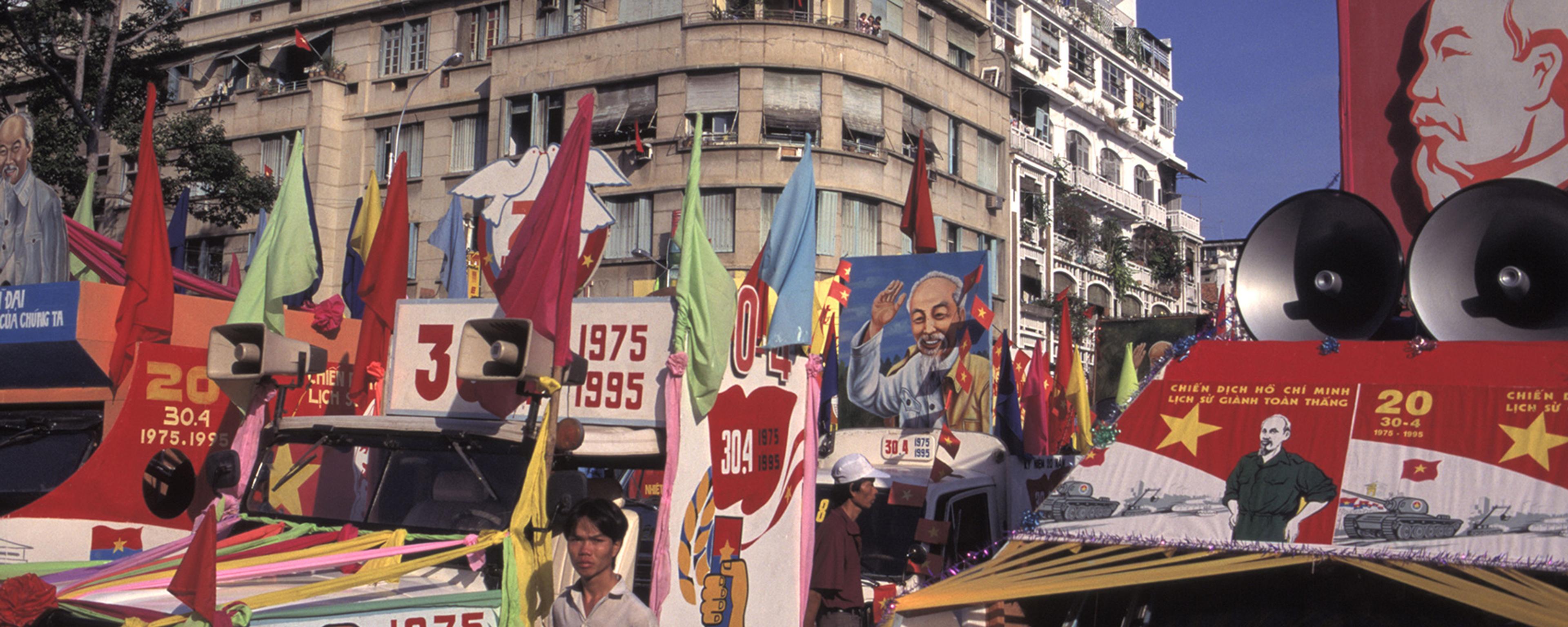 A vibrant street scene with decorated vehicles, colourful flags and banners in front of a large urban building.