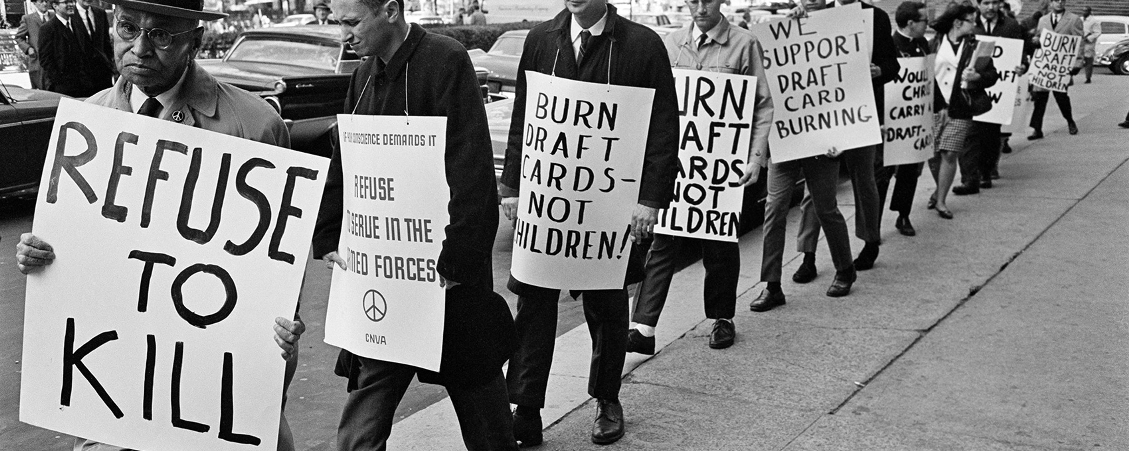 Black and white photo of a line of people marching on a city street holding signs protesting against the draft for the Vietnam War.