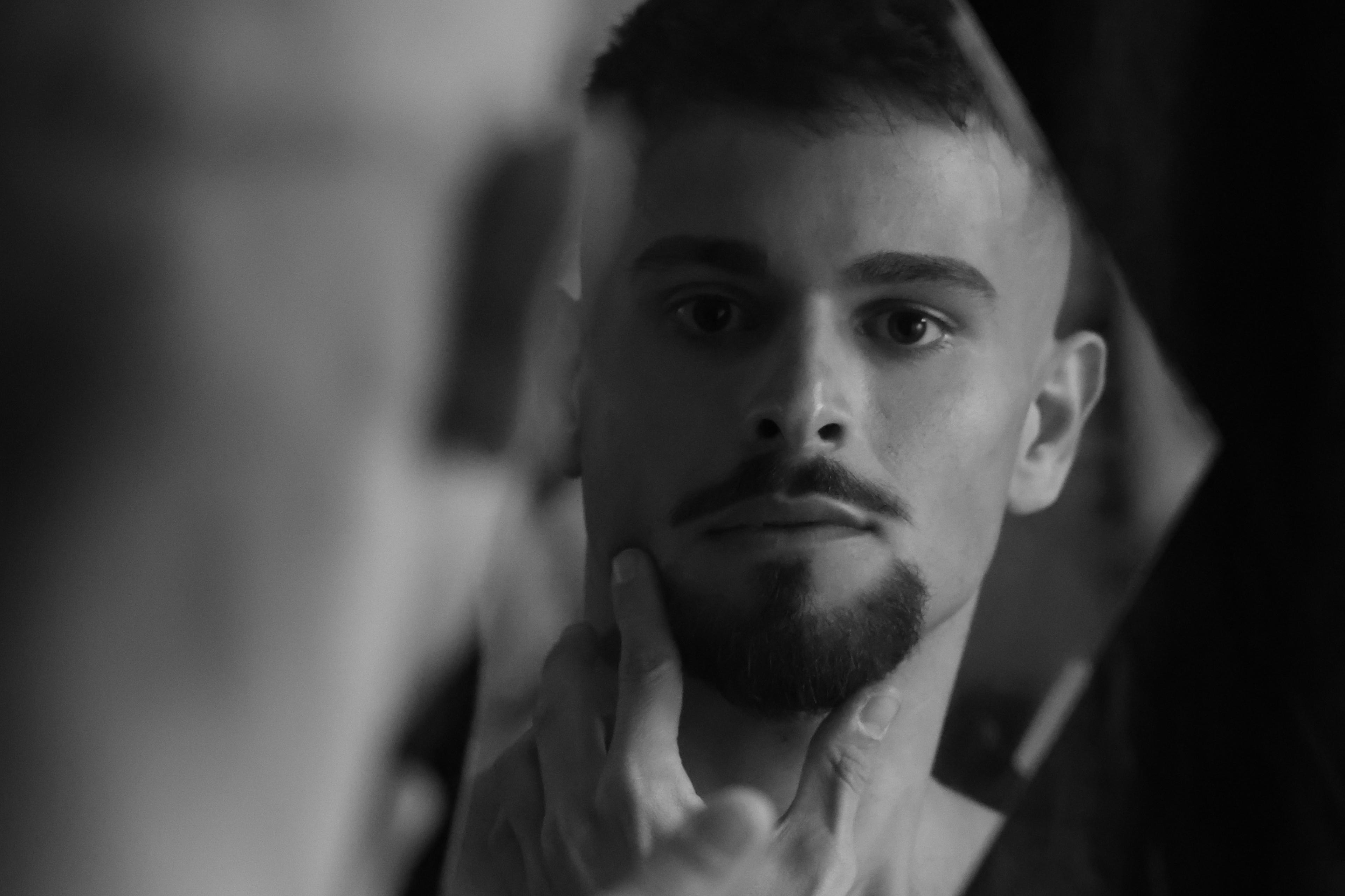 Black and white photo of a man looking in a mirror, touching his chin with his index finger.