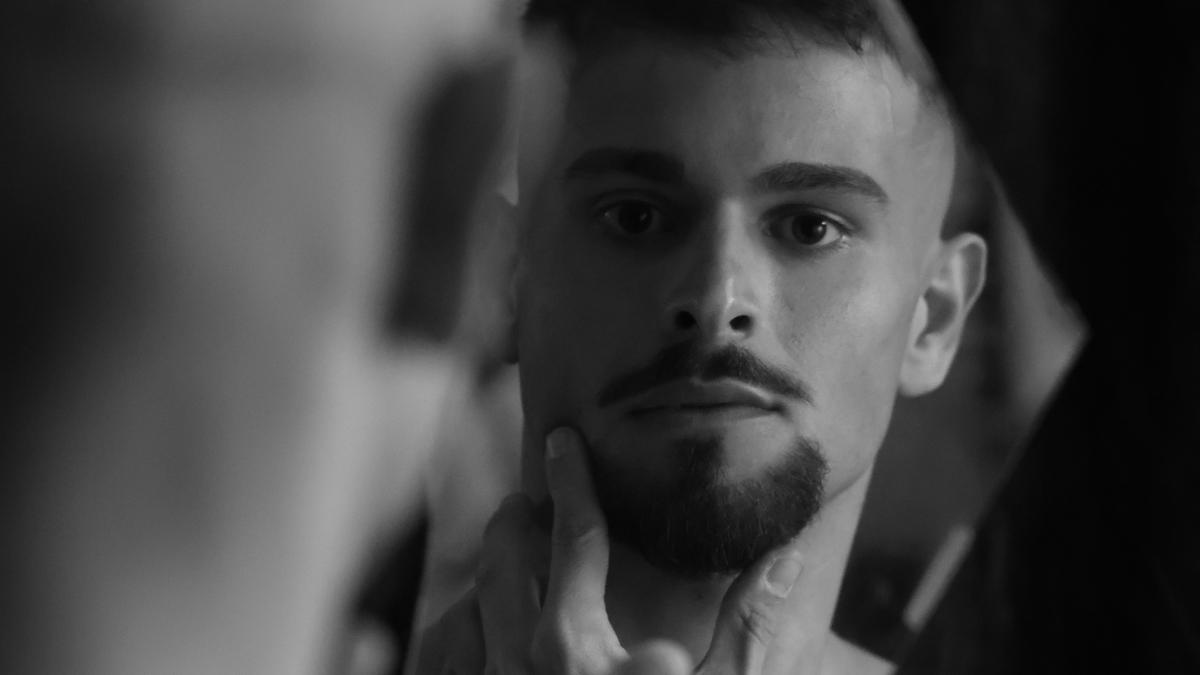Black and white photo of a man looking in a mirror, touching his chin with his index finger.
