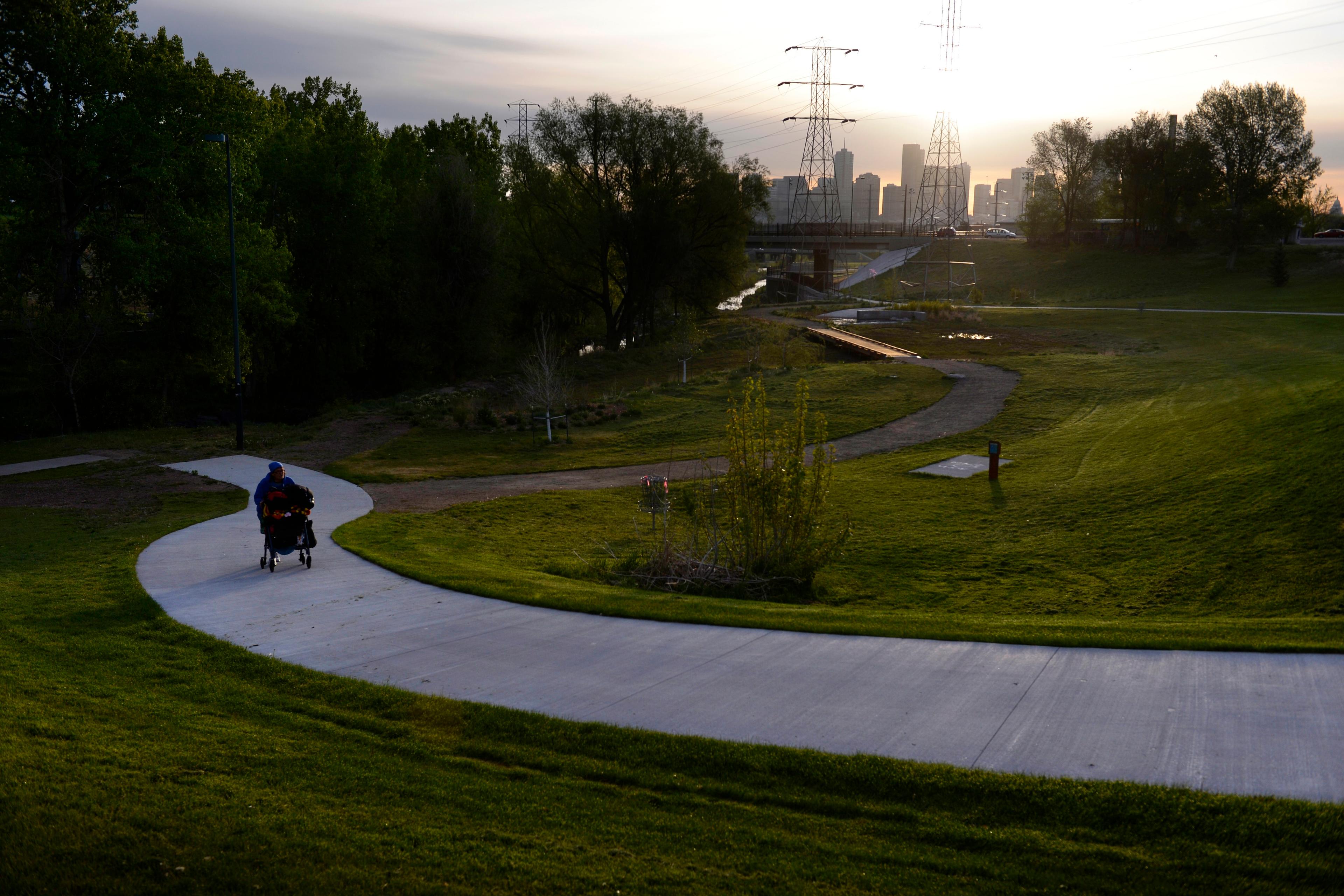 Photo of a person pushing a pram on a winding path in a park at sunset with city skyline and power lines in the background.