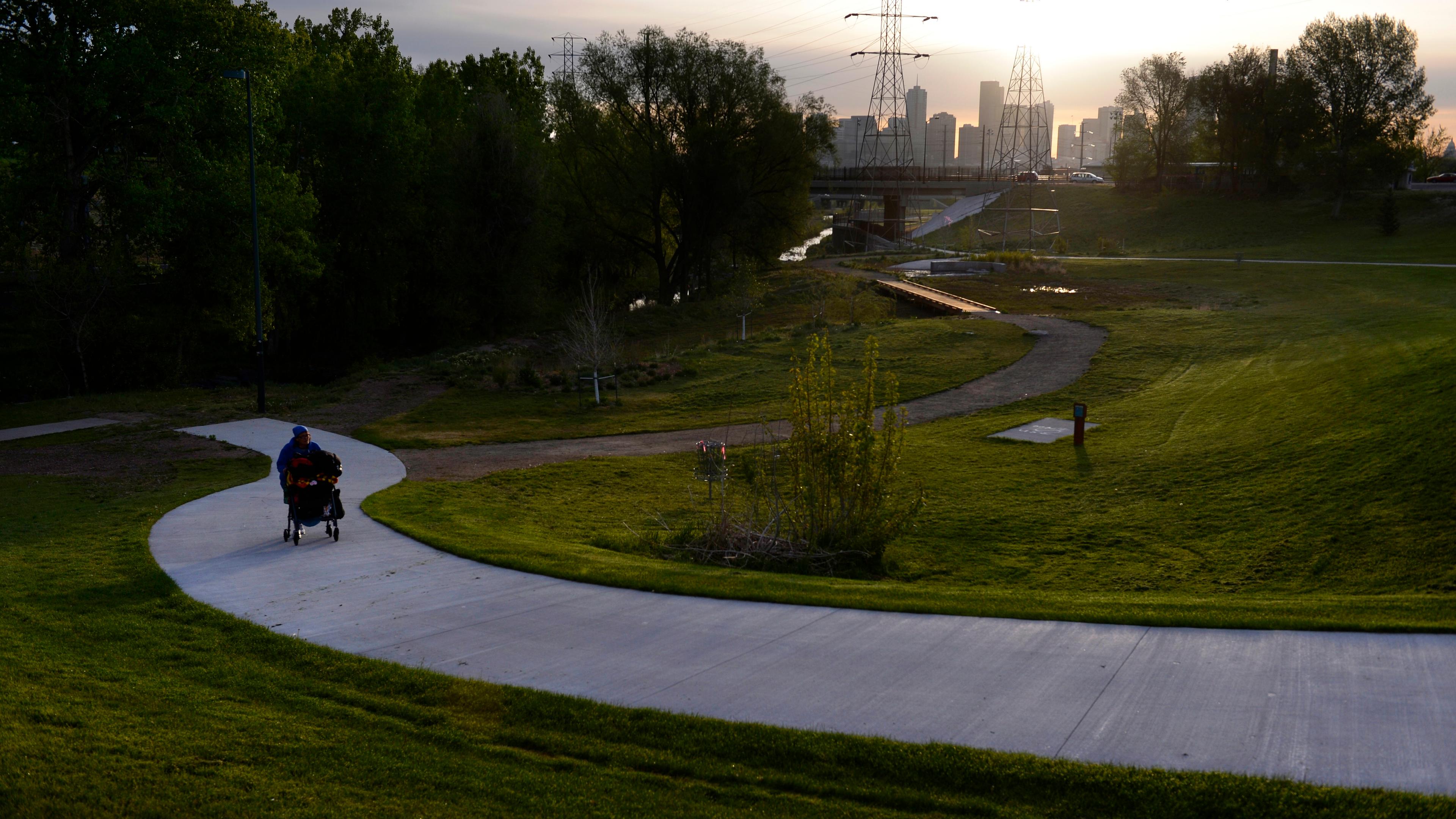 Photo of a person pushing a pram on a winding path in a park at sunset with city skyline and power lines in the background.