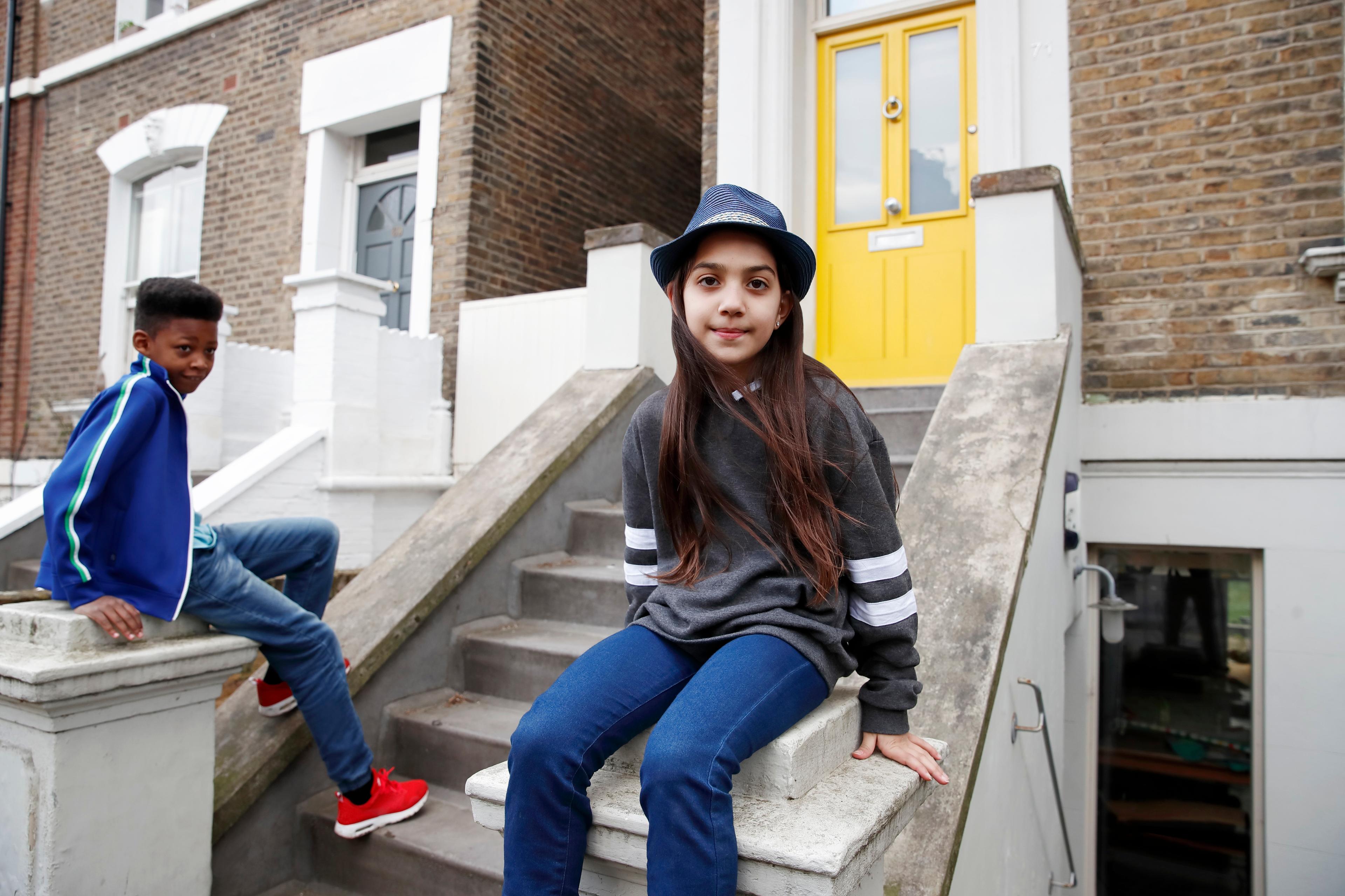 Photo of two children sitting on steps in front of a brick house with a bright yellow door, one wearing a hat.