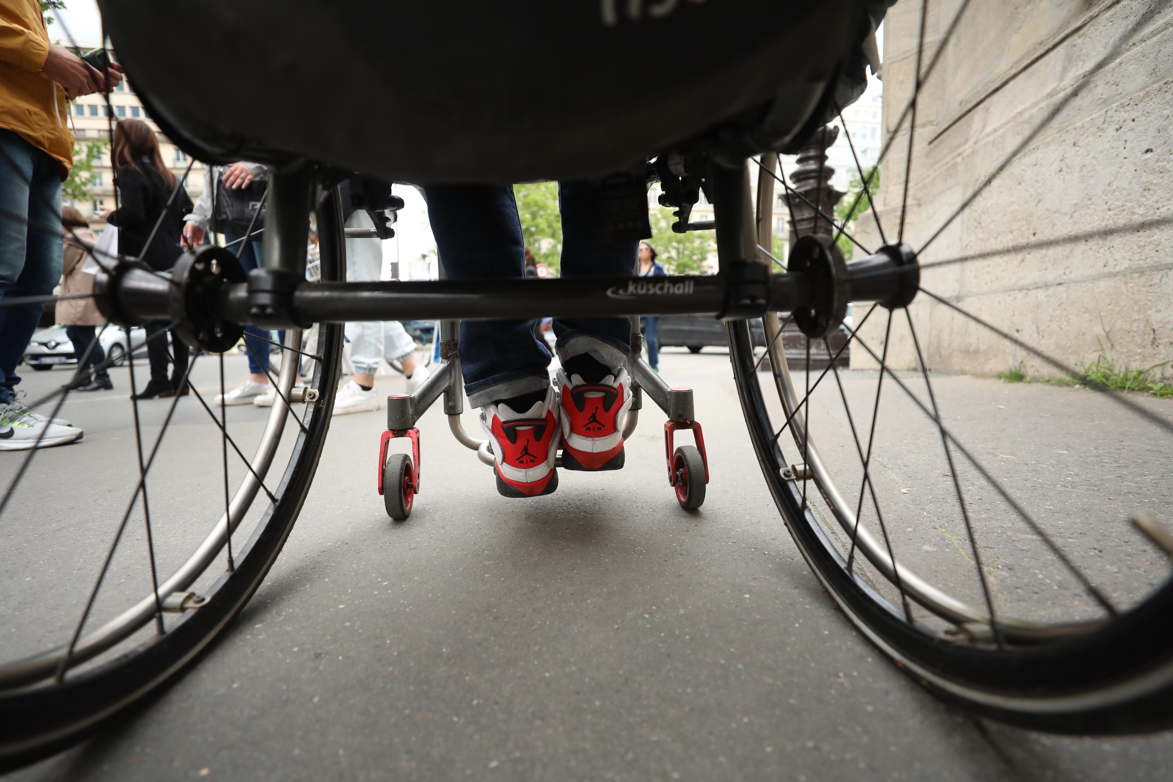 Photo of a person in a wheelchair from behind. Shoes with red details are visible. Pedestrians walk nearby on a city street.