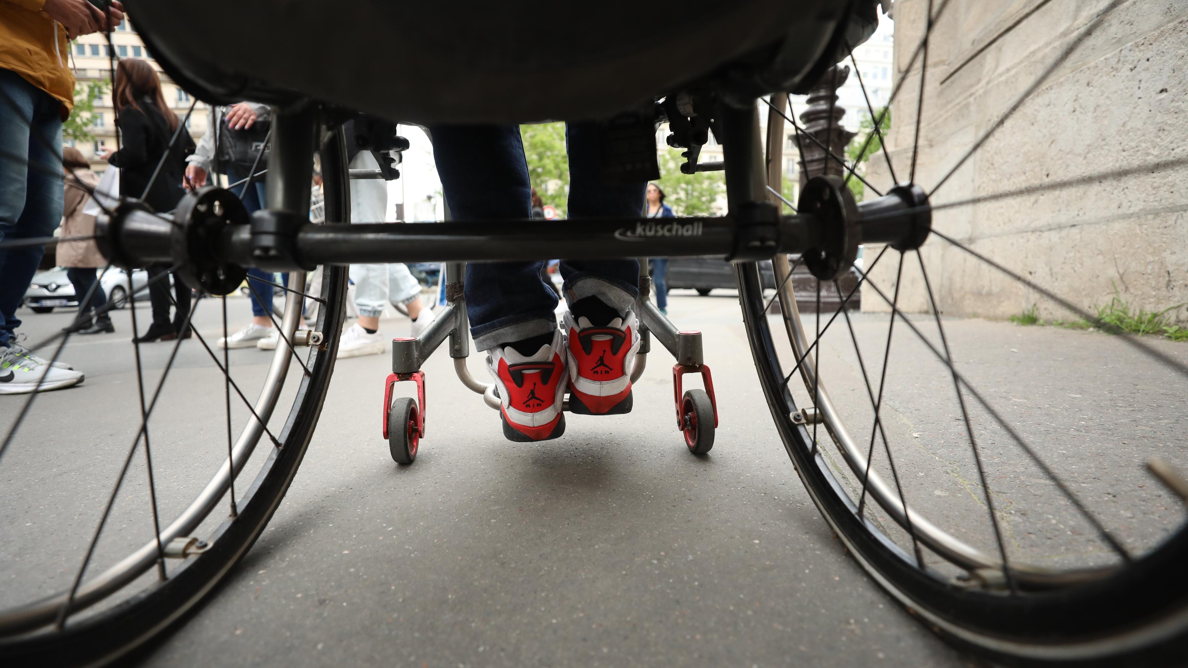 Photo of a person in a wheelchair from behind. Shoes with red details are visible. Pedestrians walk nearby on a city street.