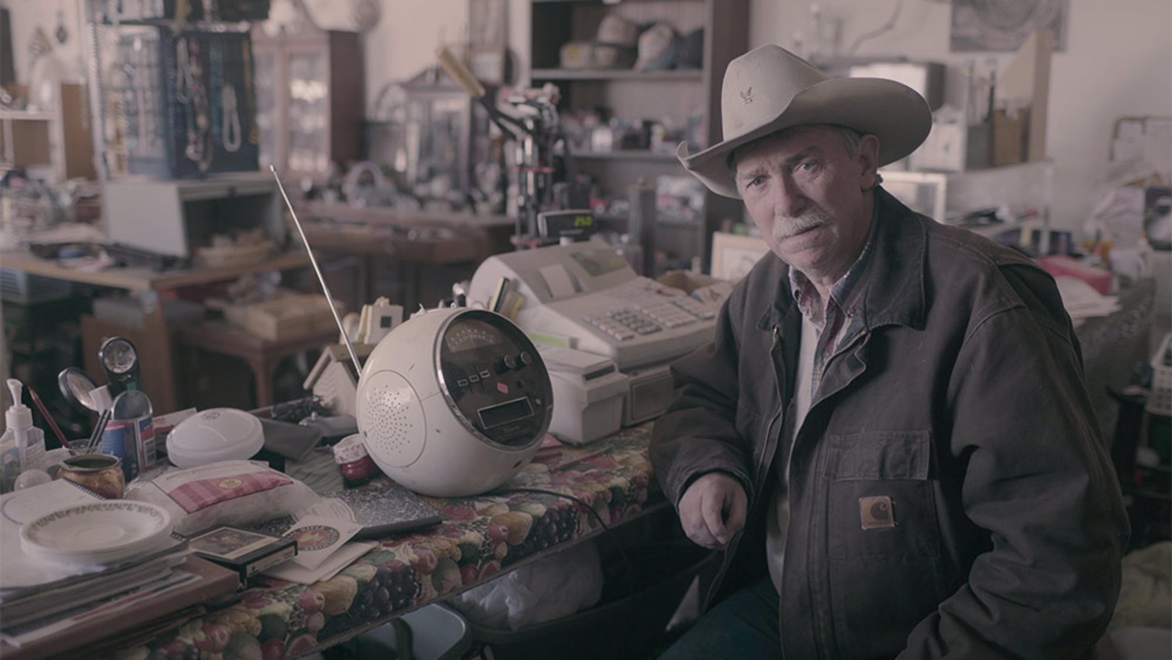 An older man in a cowboy hat and jacket sitting by a table with retro items in a cluttered antique shop.