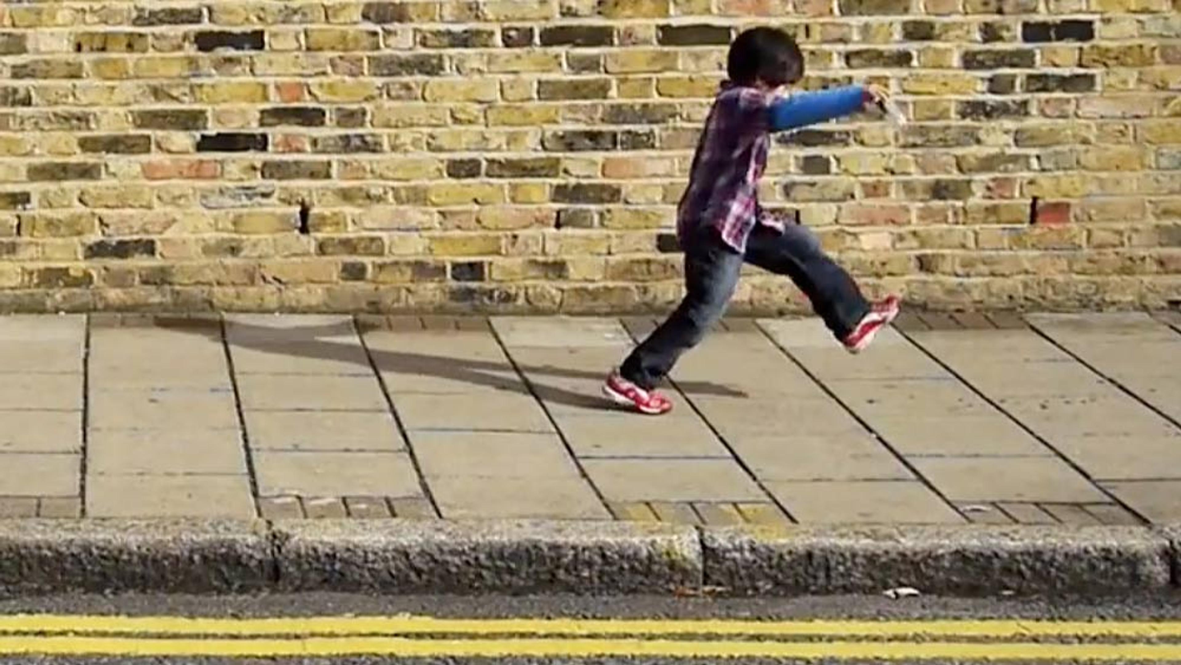 A child in a plaid shirt and jeans skipping on a pavement next to a brick wall with double yellow lines on the road.