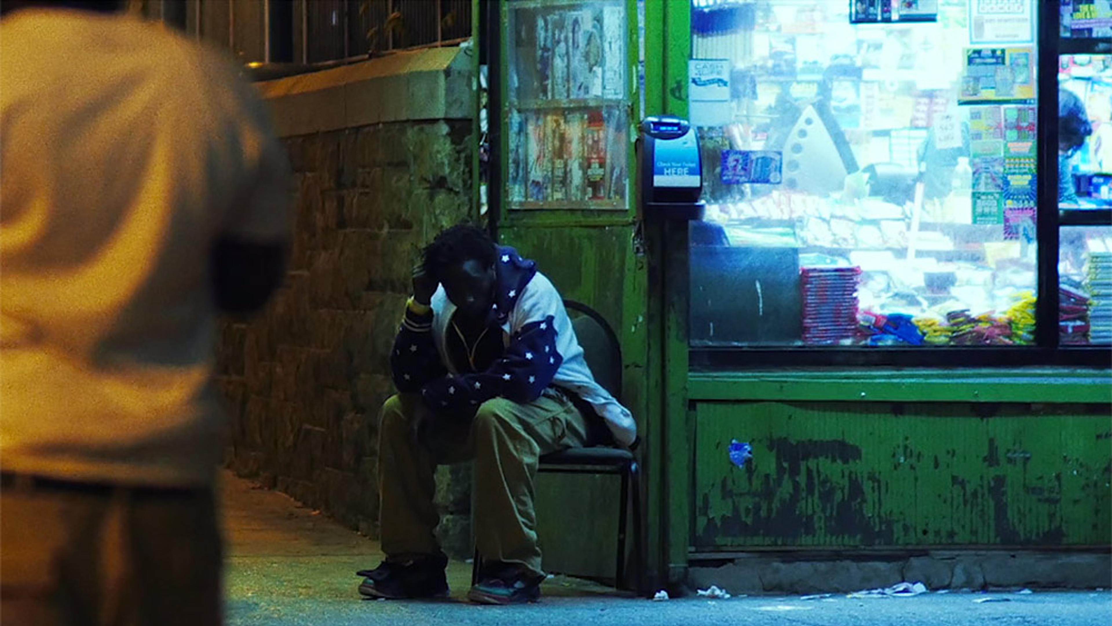 A person sitting outside a shop at night. The shop has a bright interior with many items on display.