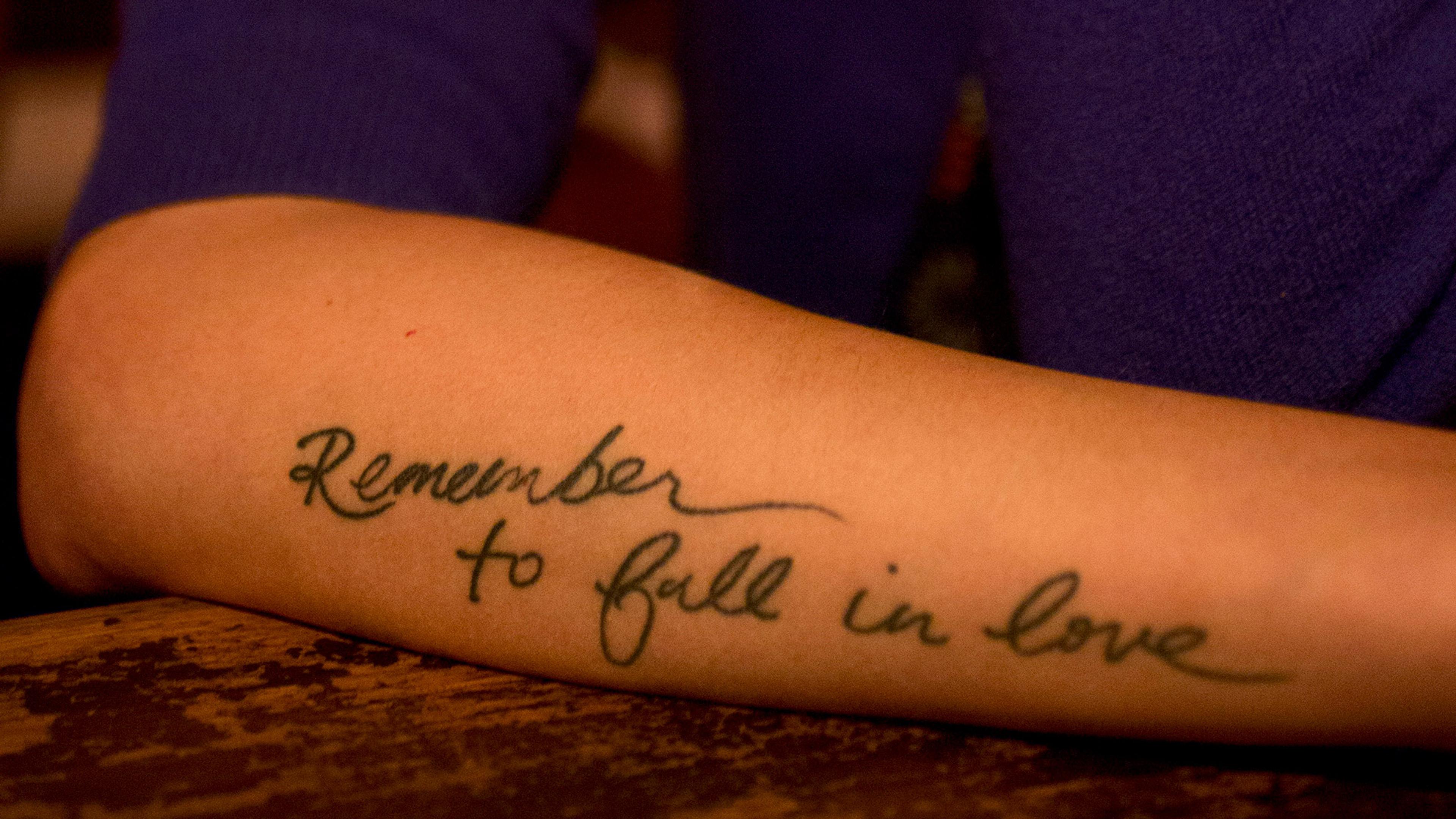 Forearm tattoo in cursive script reads “Remember to fall in love” in black ink.