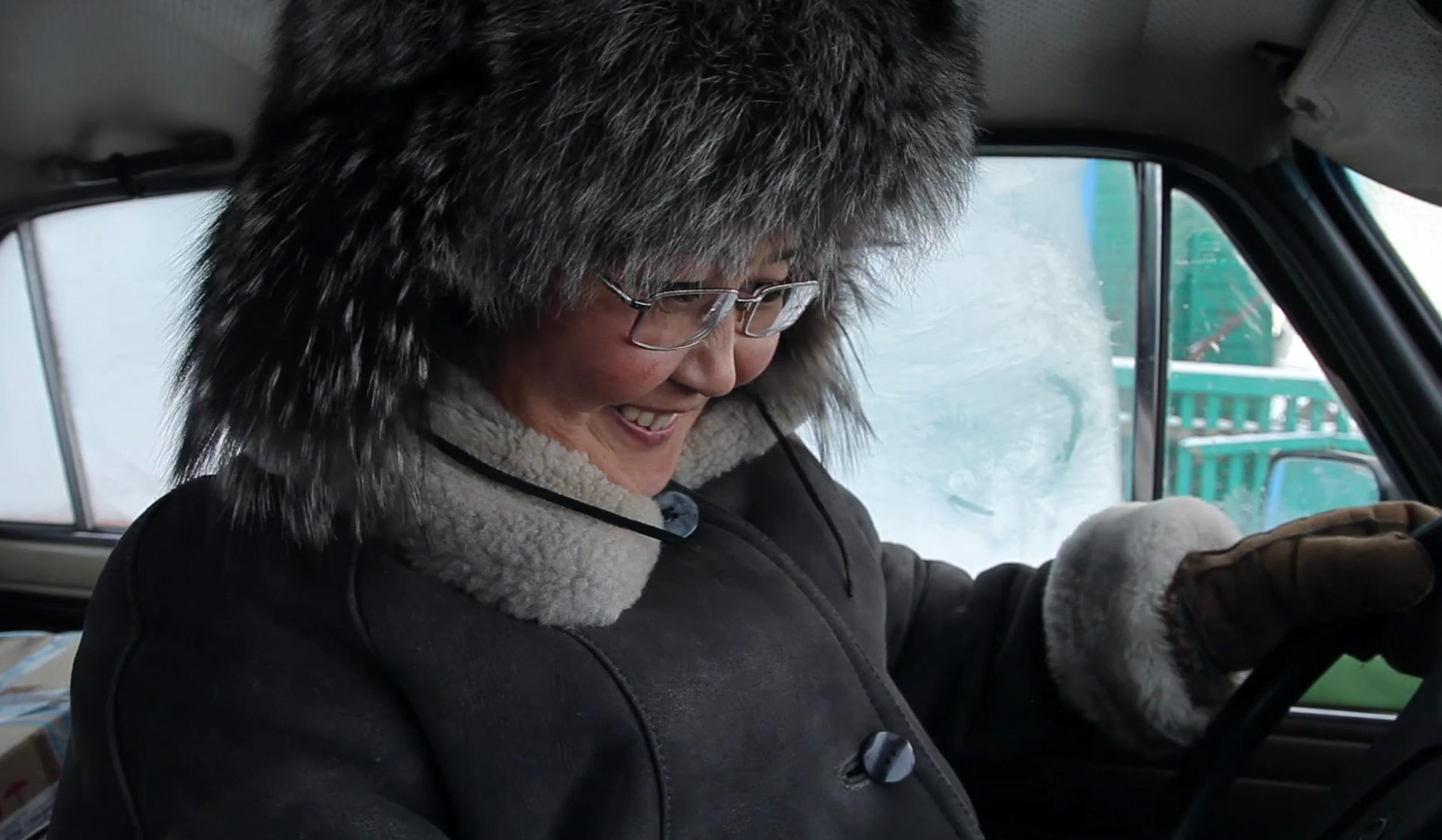 Woman in a fur hat, glasses and coat, smiling inside a vehicle with frosted windows.