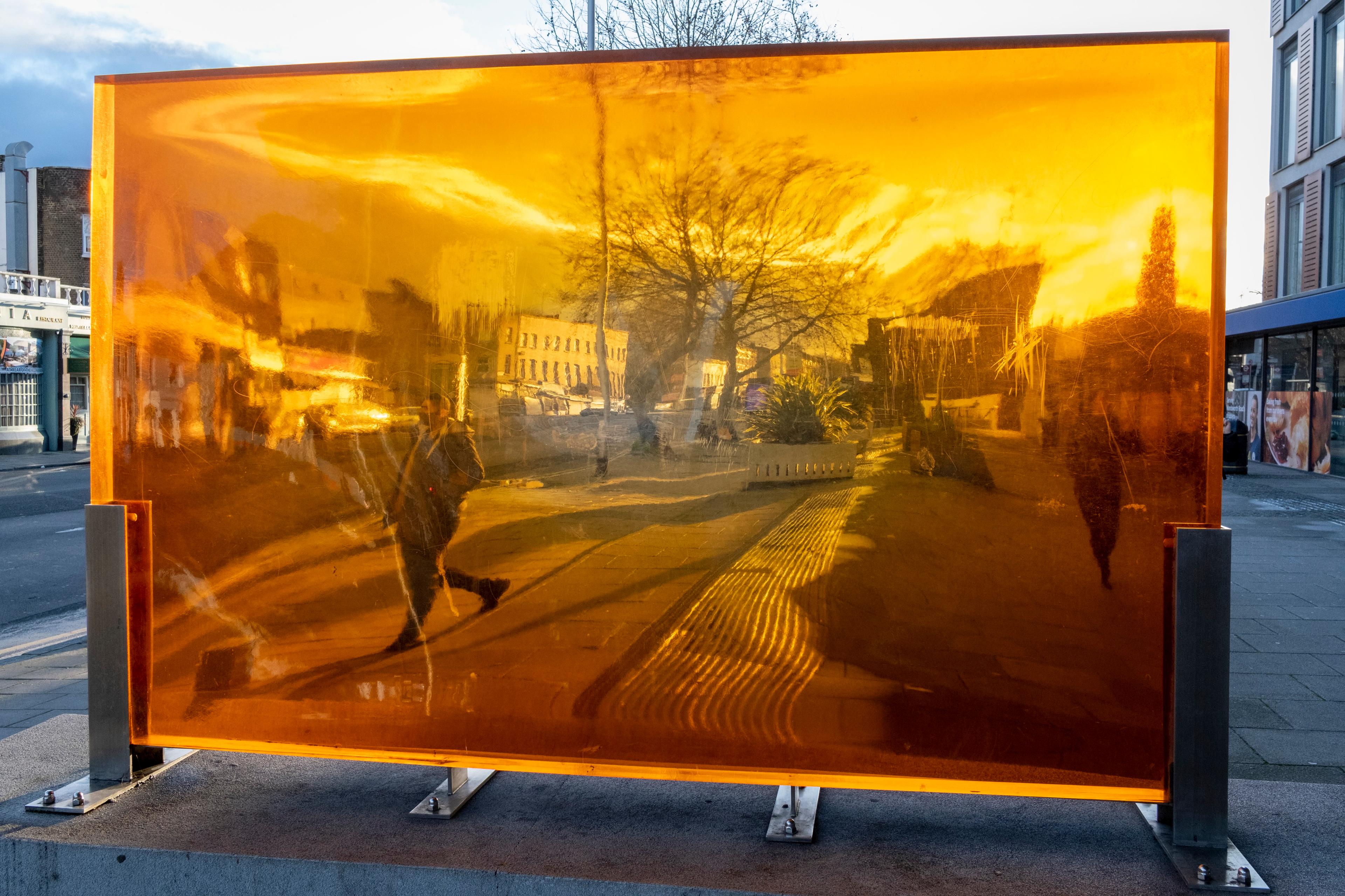 Photo of a street scene reflected in a large orange-tinted glass pane with buildings and trees distorted by the surface.