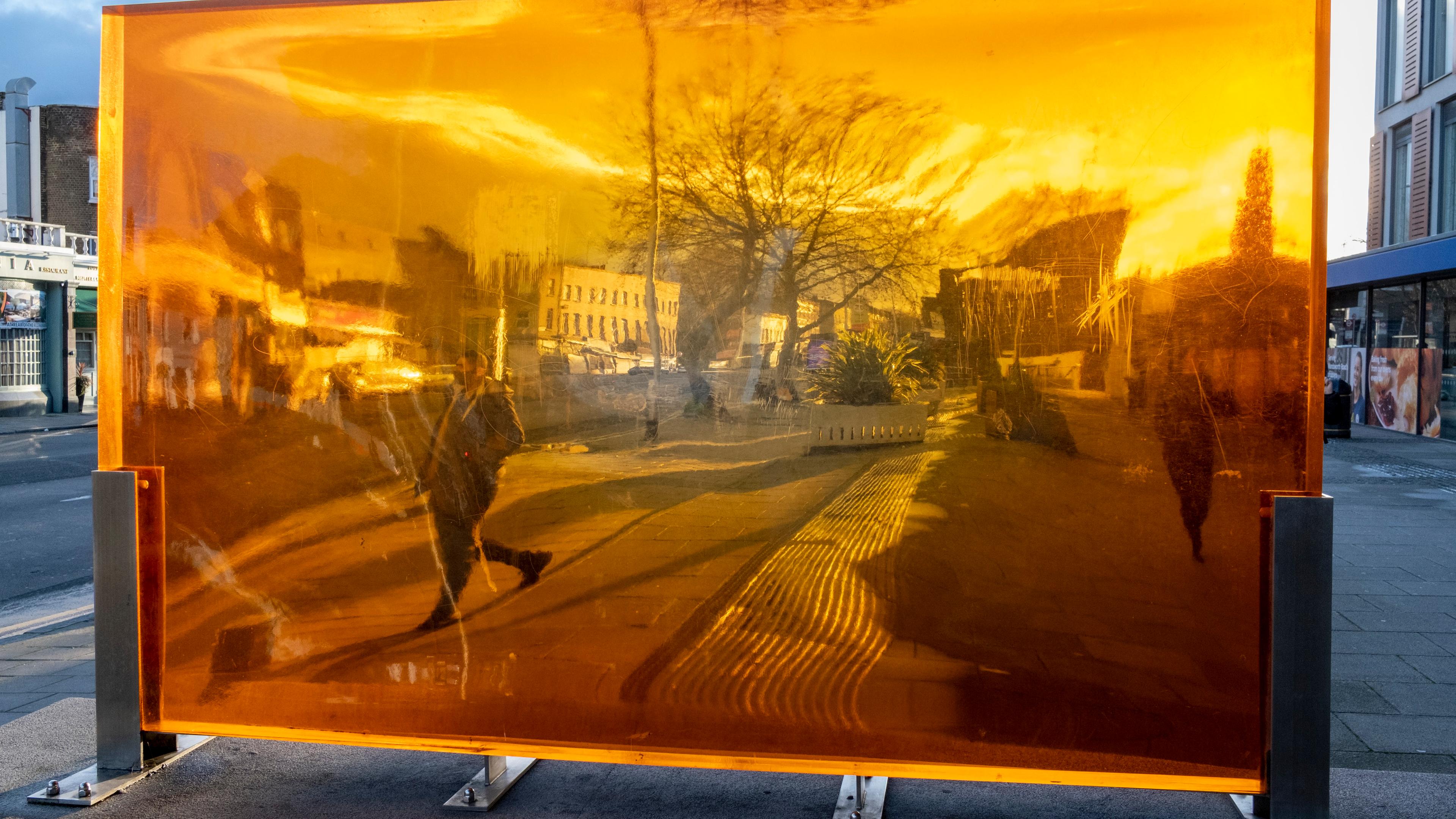 Photo of a street scene reflected in a large orange-tinted glass pane with buildings and trees distorted by the surface.