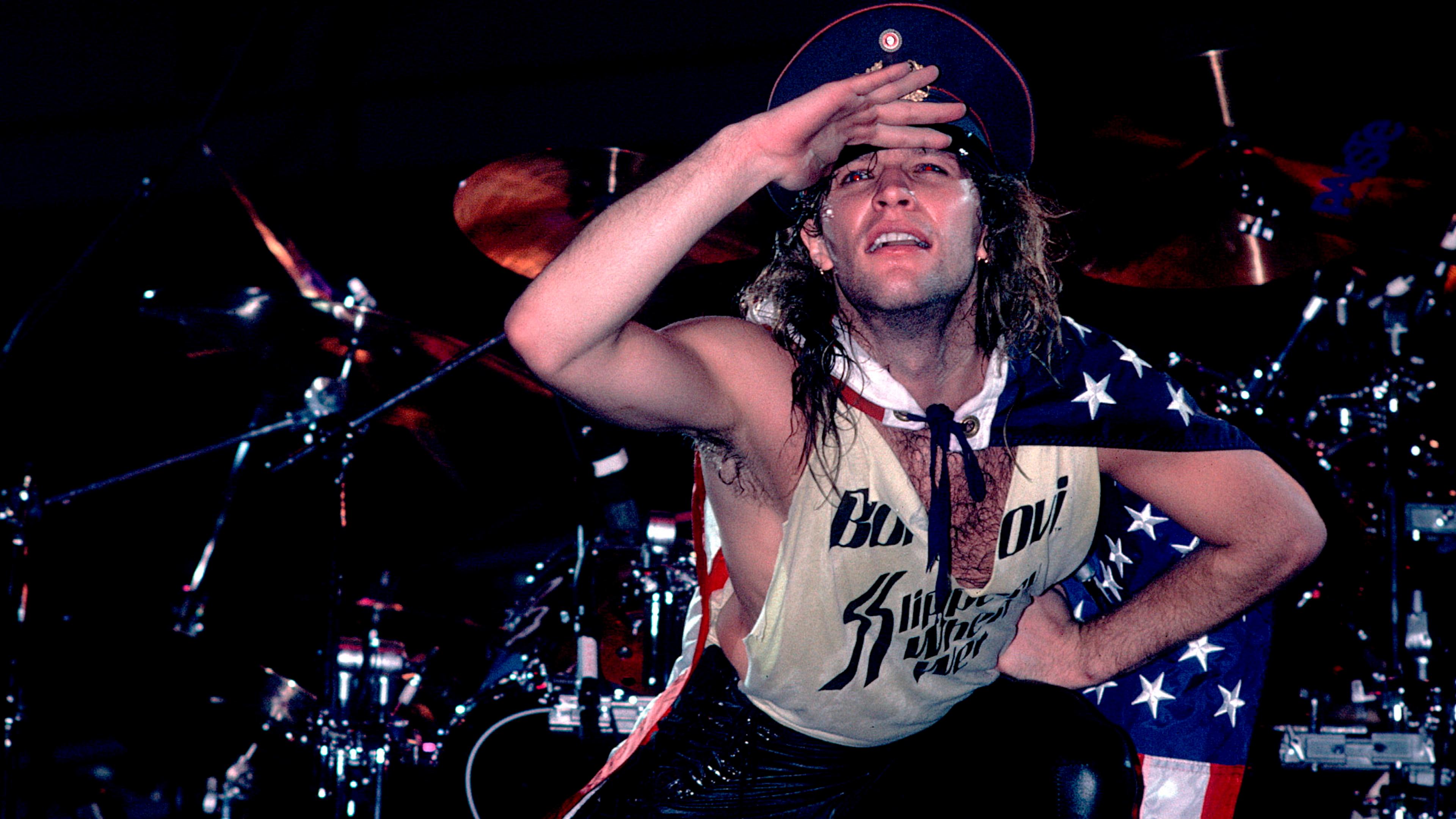 Photo of a performer on stage wearing a vest and hat, posing energetically with a US flag draped on their back.