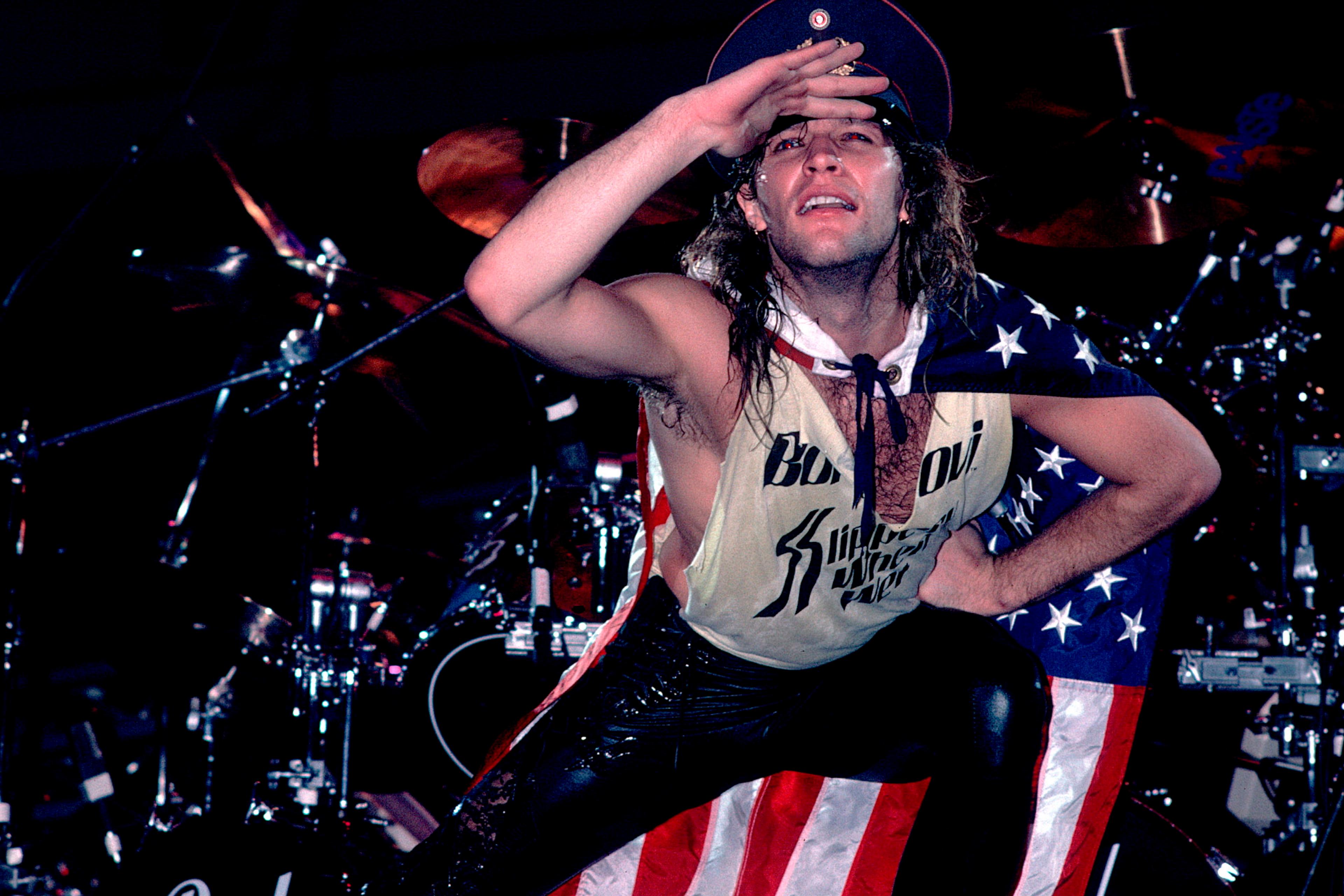 Photo of a performer on stage wearing a vest and hat, posing energetically with a US flag draped on their back.