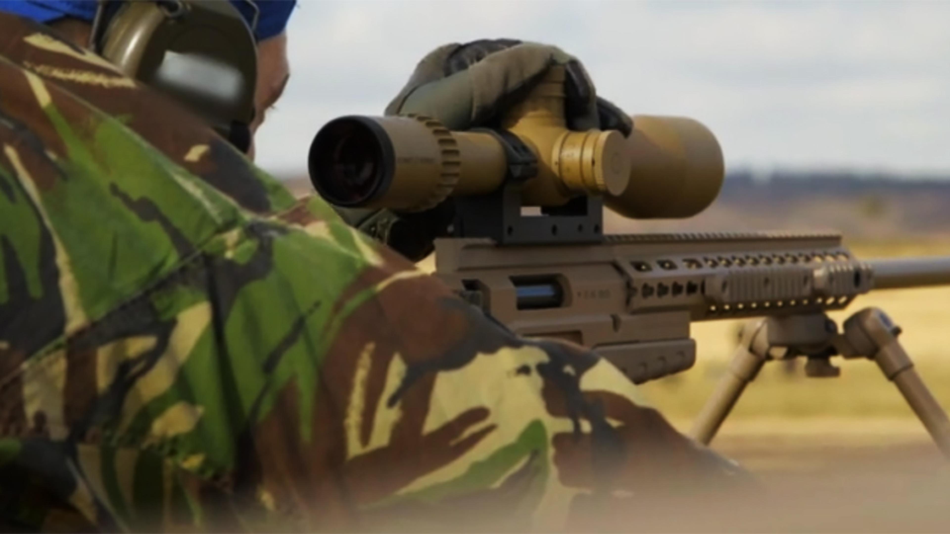 A person in camouflage aiming a rifle with a scope while lying prone outdoors, wearing ear defenders.