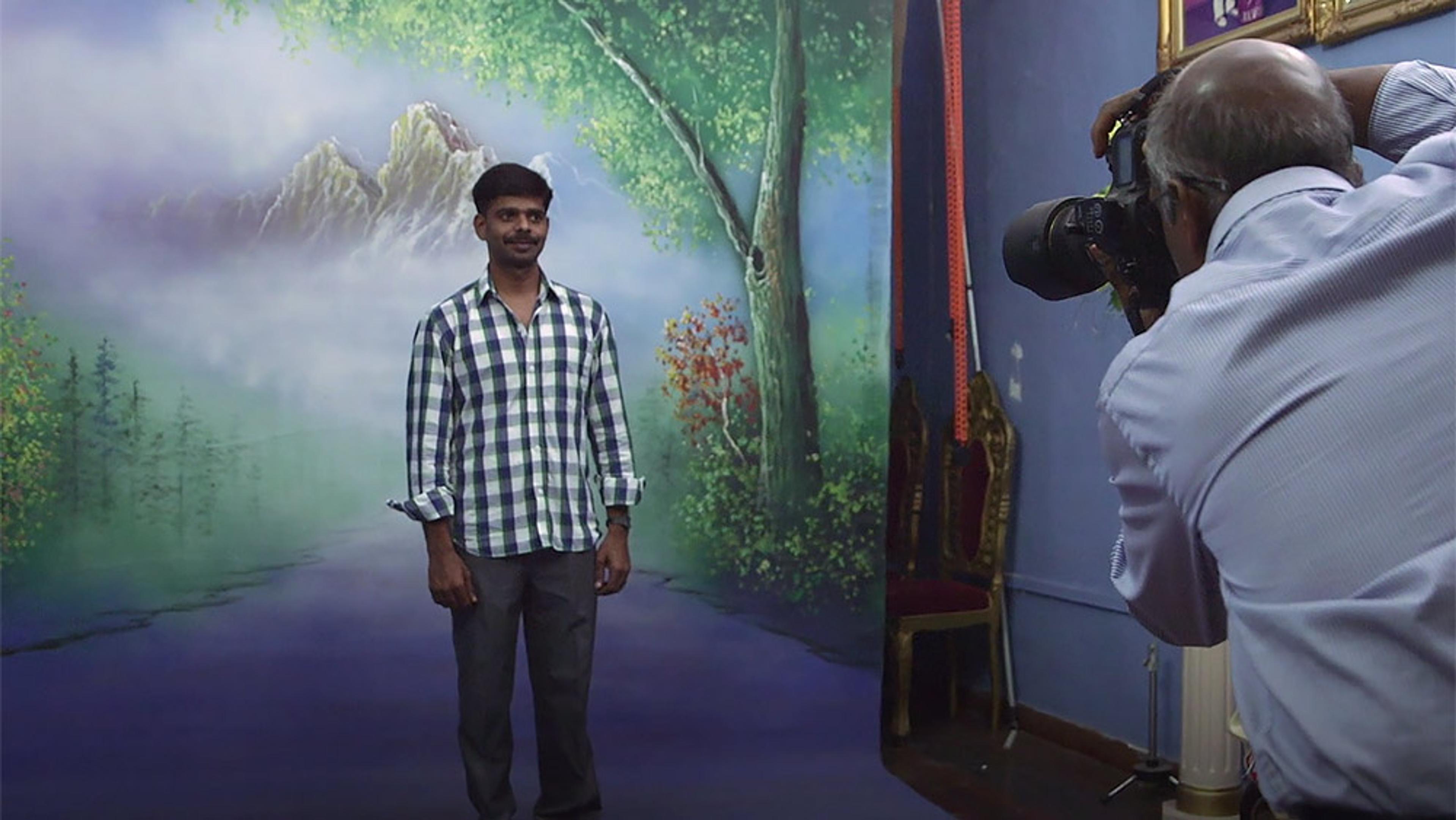 A man posing in front of a scenic backdrop while a photographer takes his picture in a studio setting.