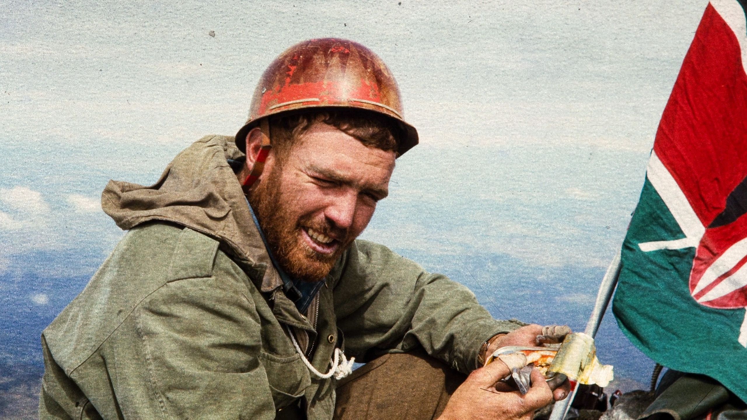 Photo of a man in a helmet and jacket eating a snack next to a flag on a mountain summit under a clear sky.