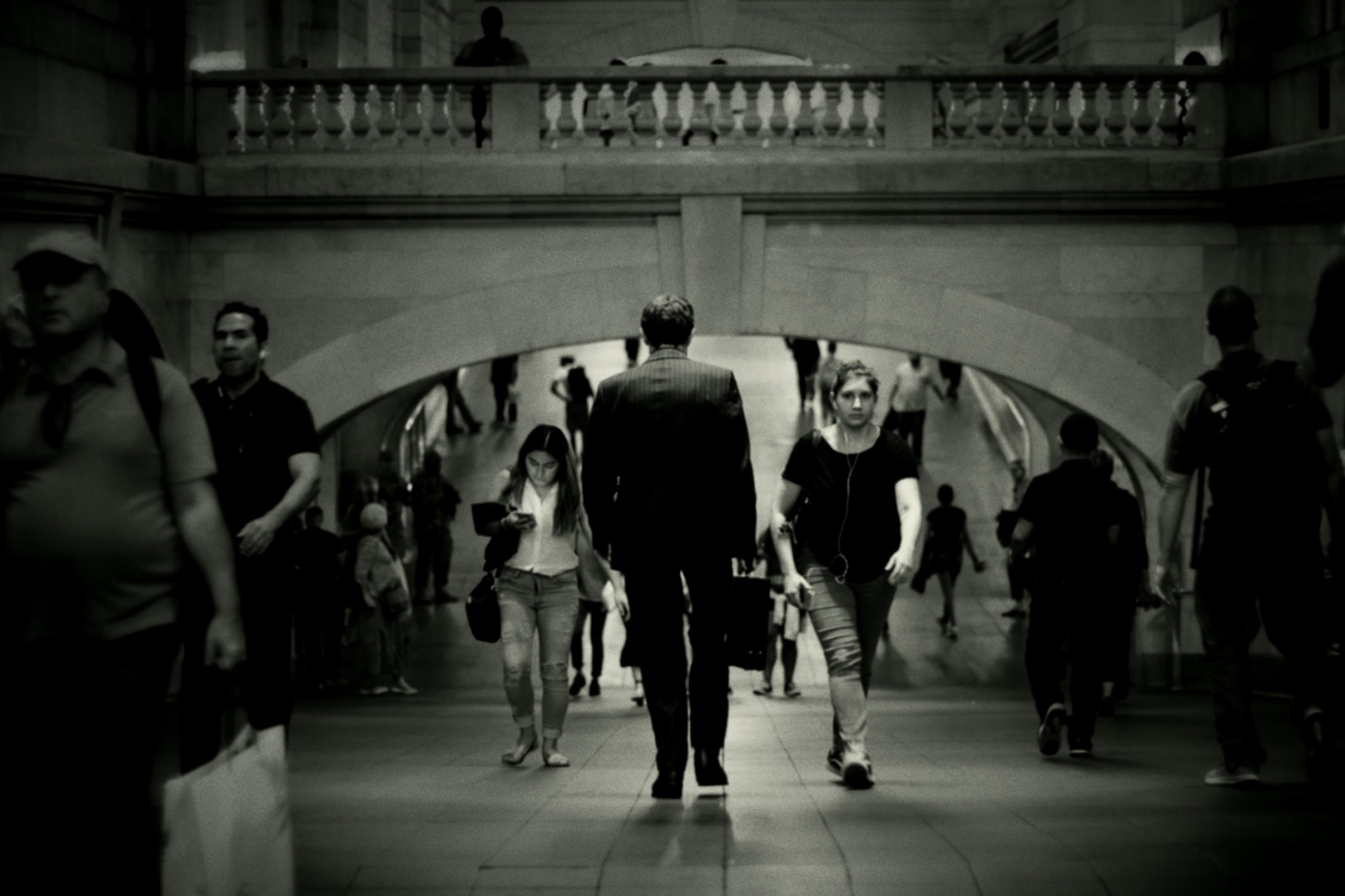 Black-and-white image of people walking in a busy station corridor with an arched bridge above them.