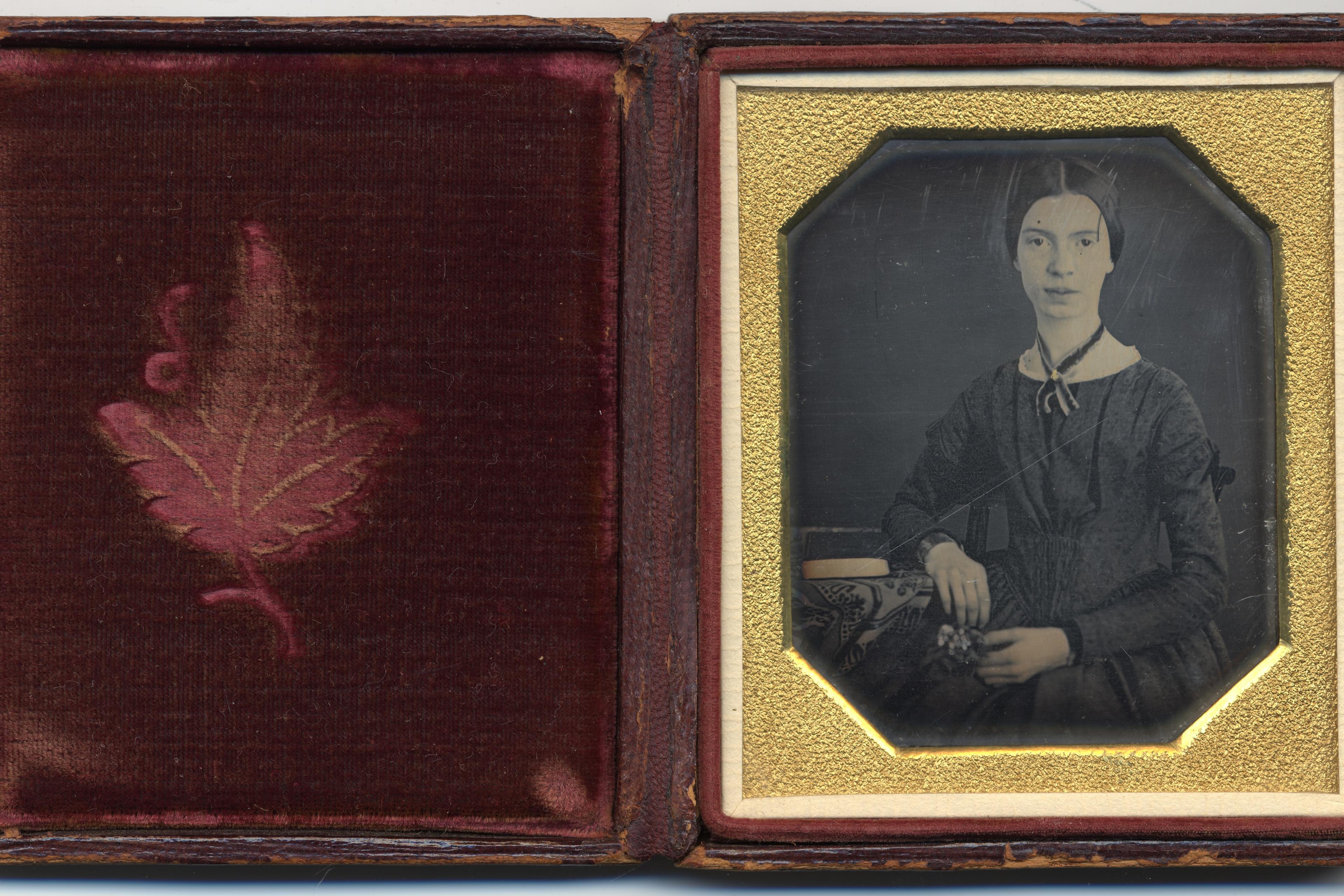 Daguerreotype of a woman in a dark dress seated with hands on her lap in a gold frame next to a red velvet case with a leaf design.