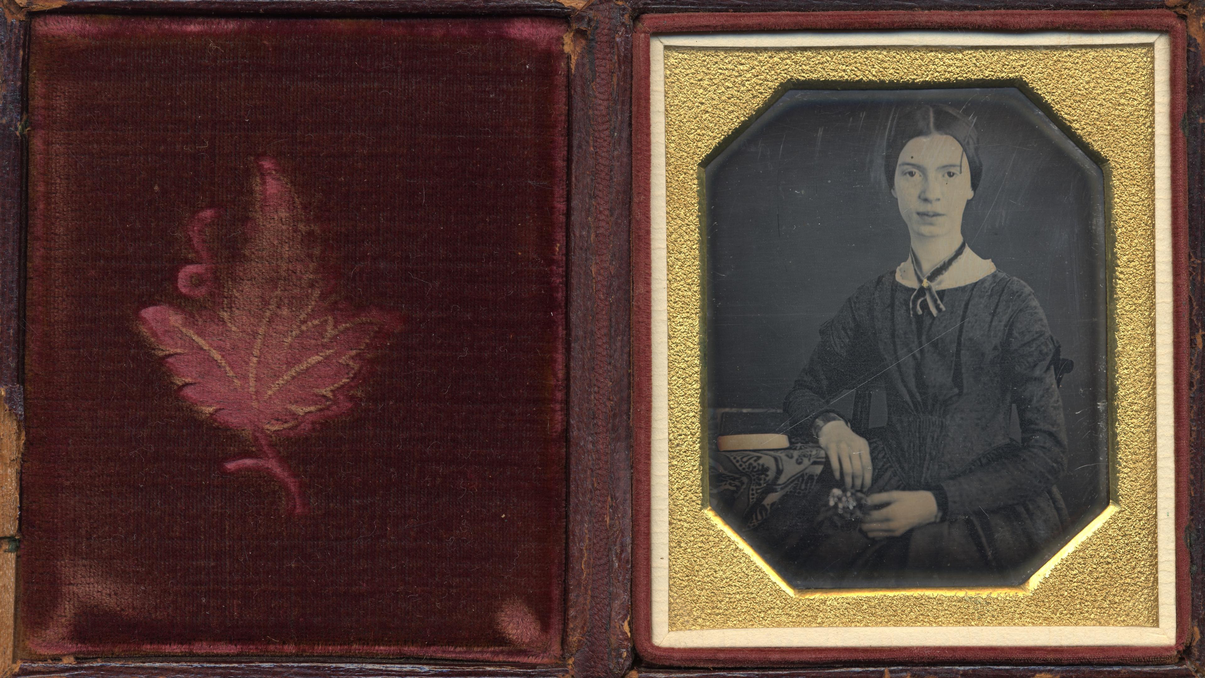 Daguerreotype of a woman in a dark dress seated with hands on her lap in a gold frame next to a red velvet case with a leaf design.