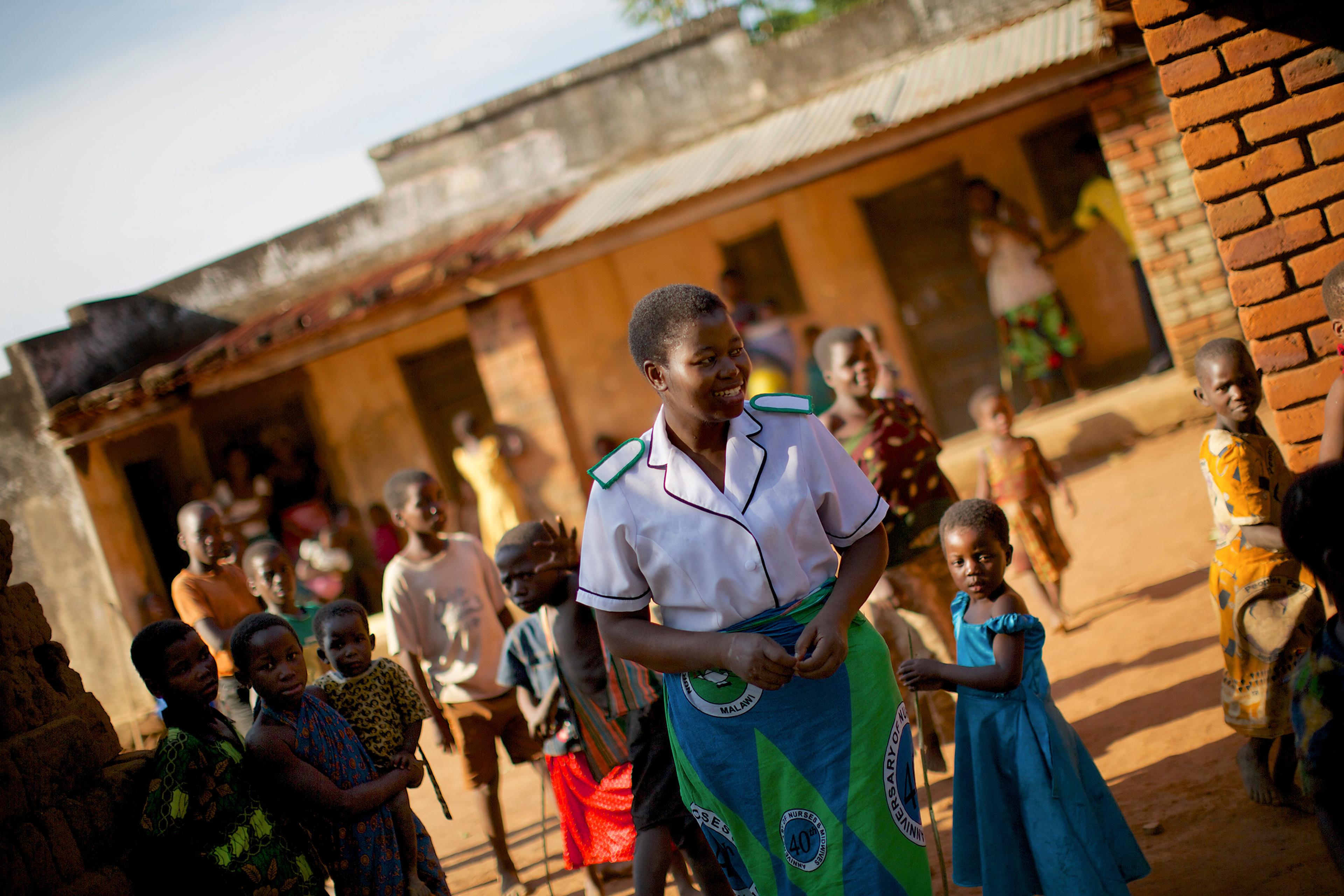 A smiling woman in a nurse’s uniform surrounded by children in an outdoor setting with brick buildings.