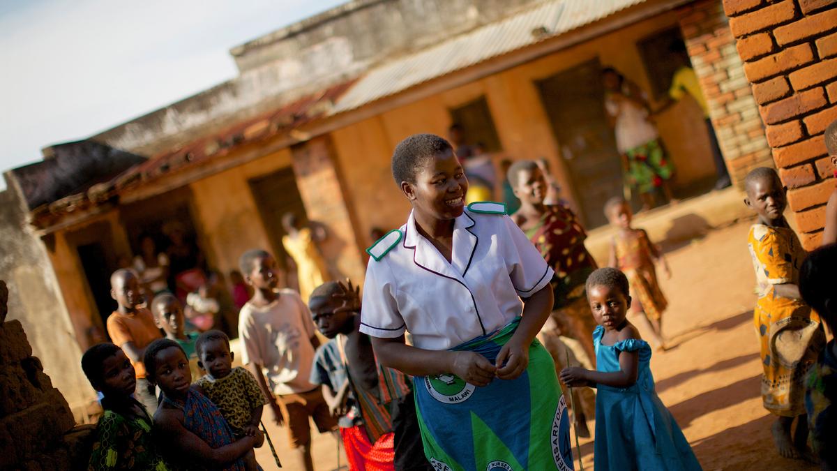 A smiling woman in a nurse’s uniform surrounded by children in an outdoor setting with brick buildings.