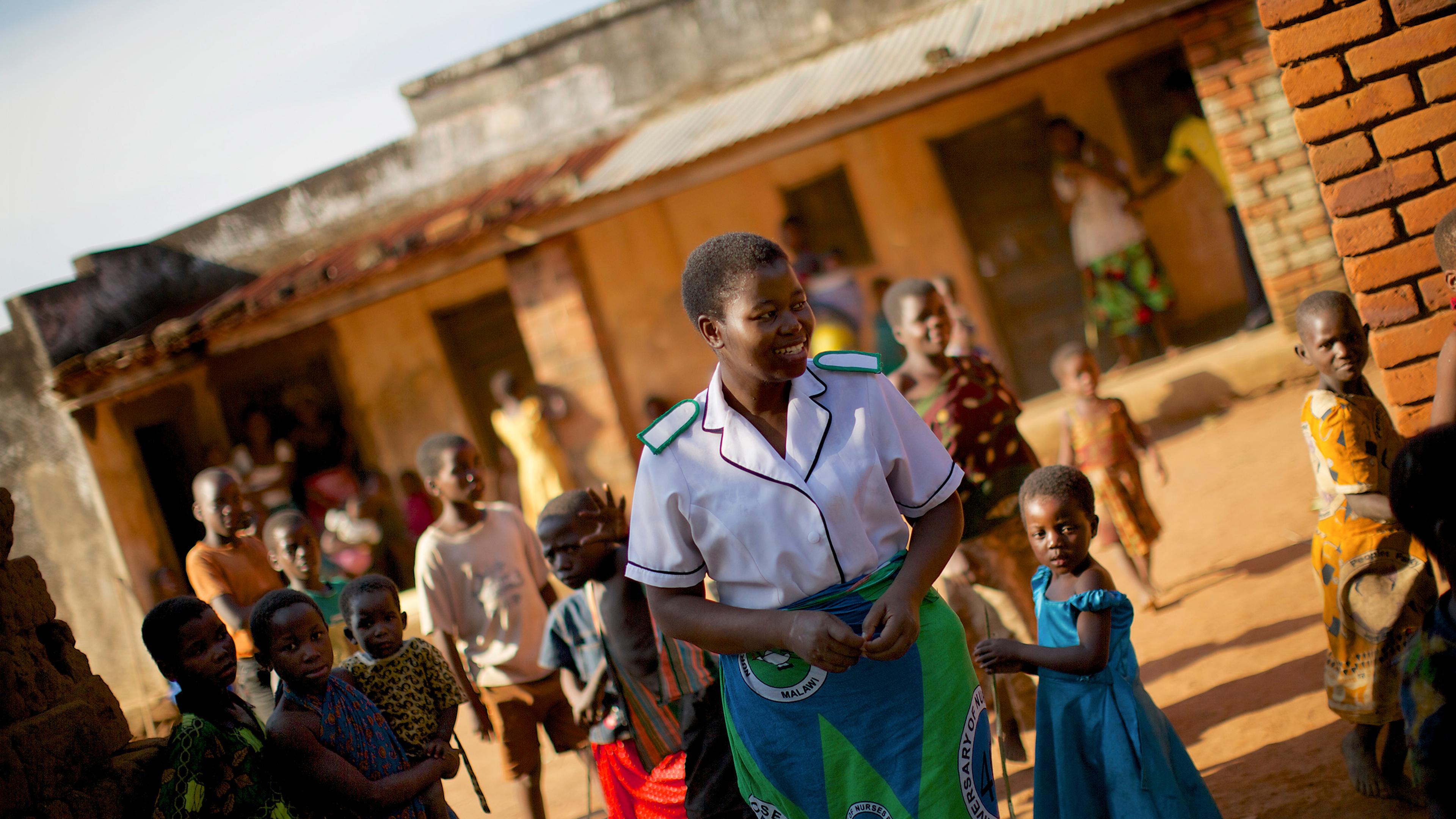 A smiling woman in a nurse’s uniform surrounded by children in an outdoor setting with brick buildings.