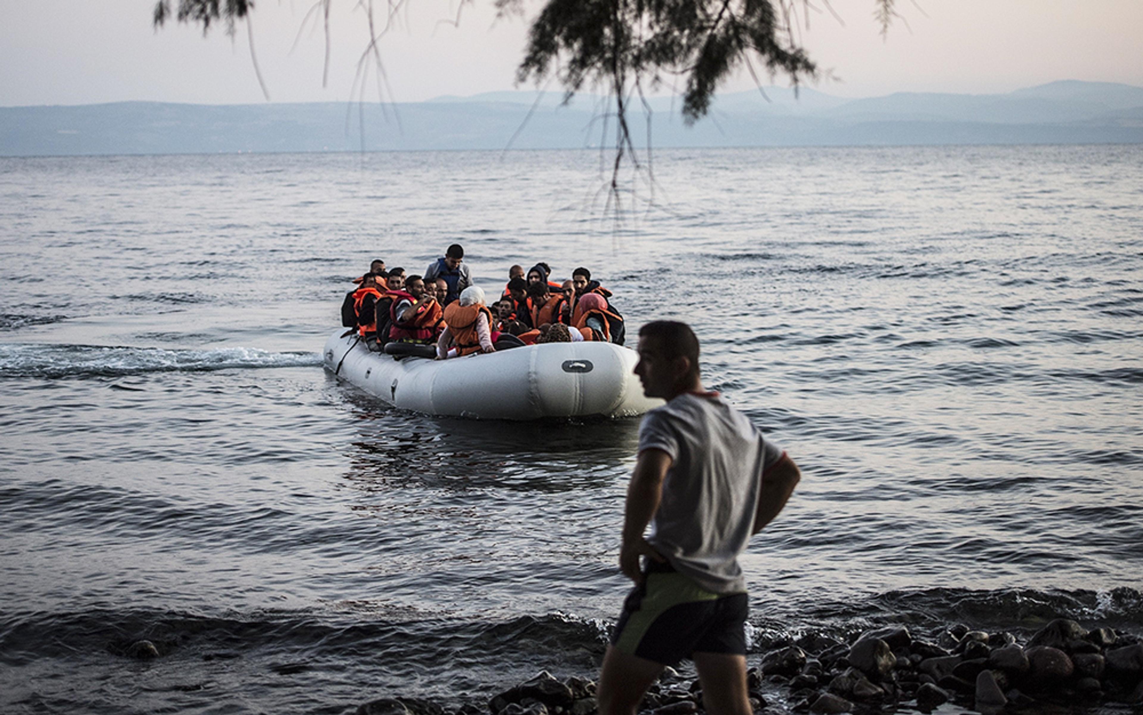 People in life jackets on a crowded inflatable boat approaching a rocky shore with a person standing on the beach.