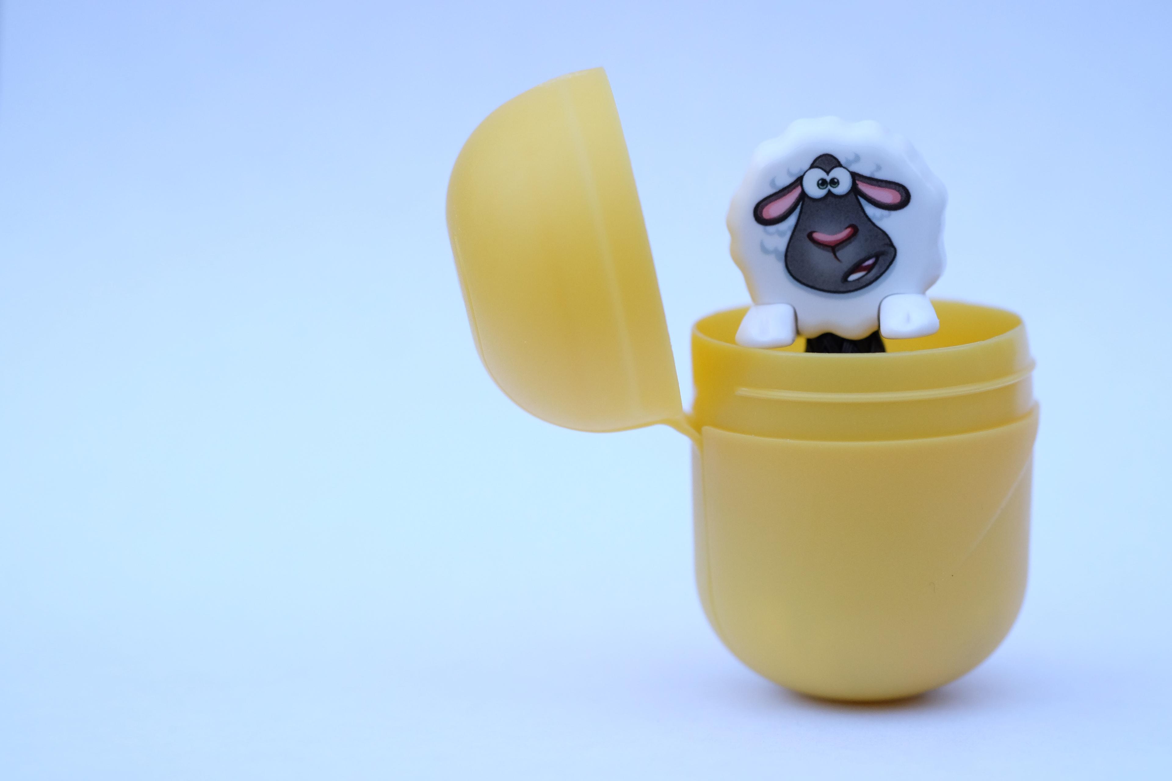 Photo of a toy sheep peeking out of an open yellow plastic capsule, set against a light blue background.