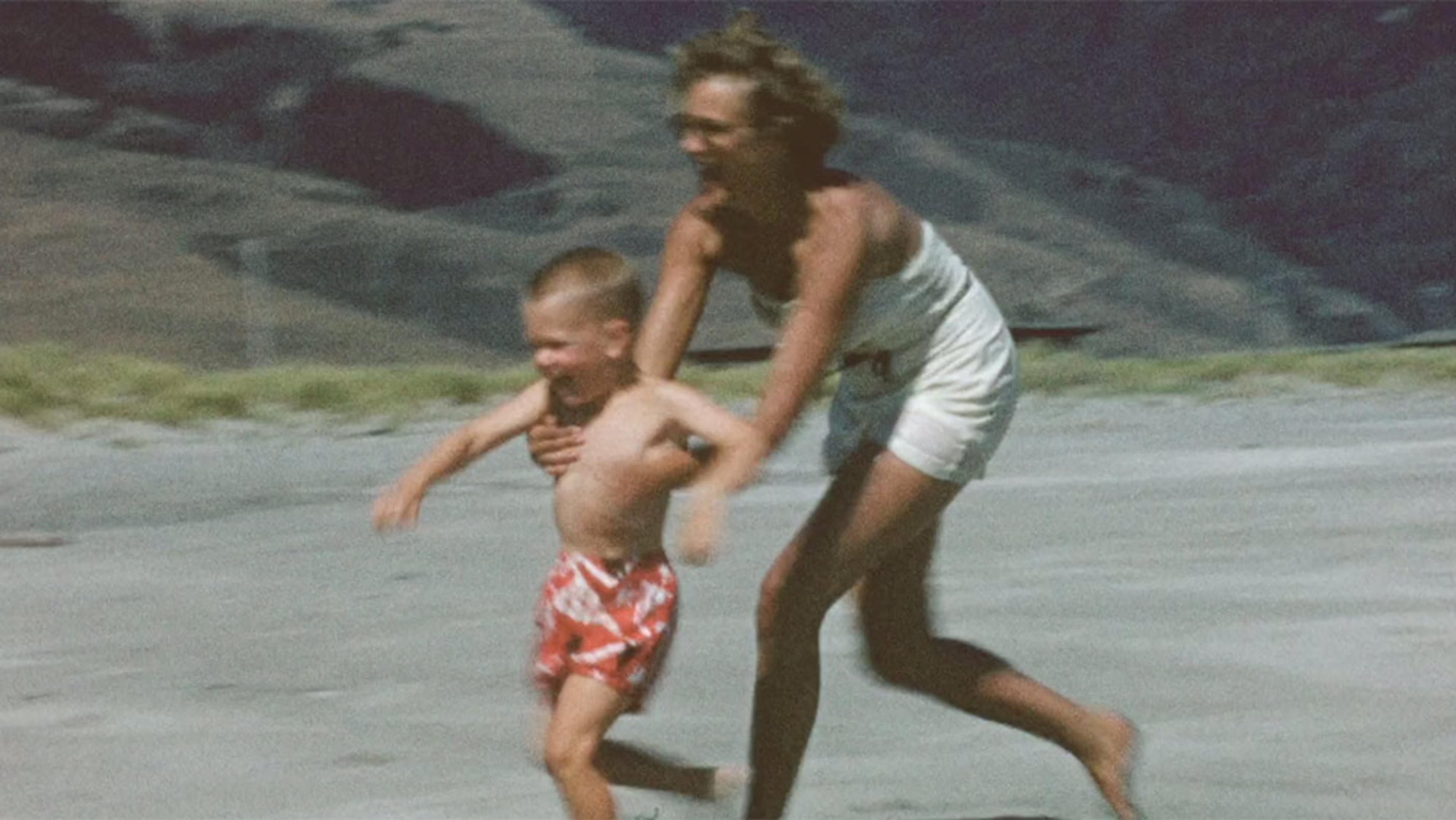 A vintage photo of a blonde woman and child running on a beach, the woman is wearing white shorts, the child wears red and white trunks.