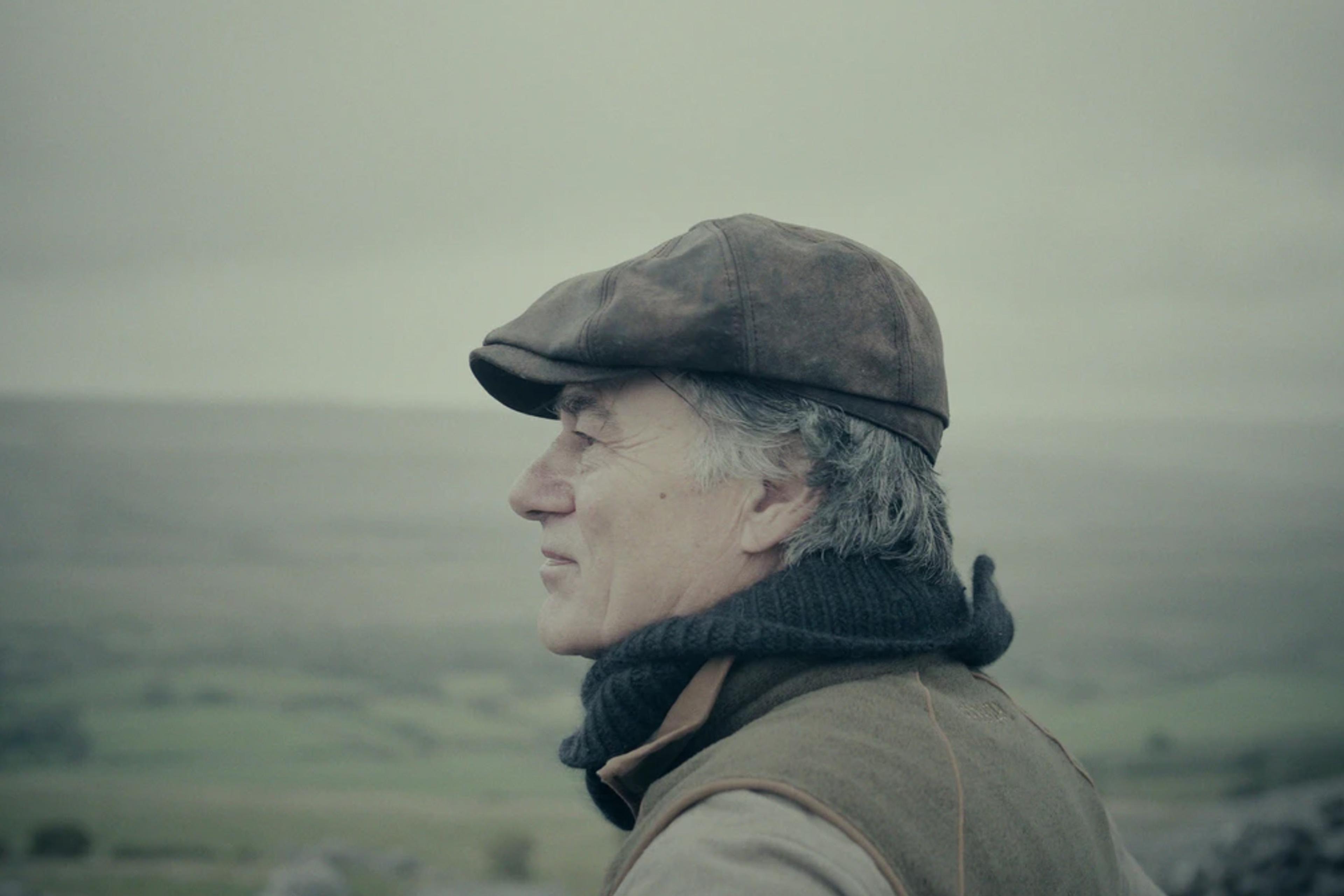 Close-up of an older man in a flat cap and scarf, looking out over a misty rural landscape with blurred farmland in the background.