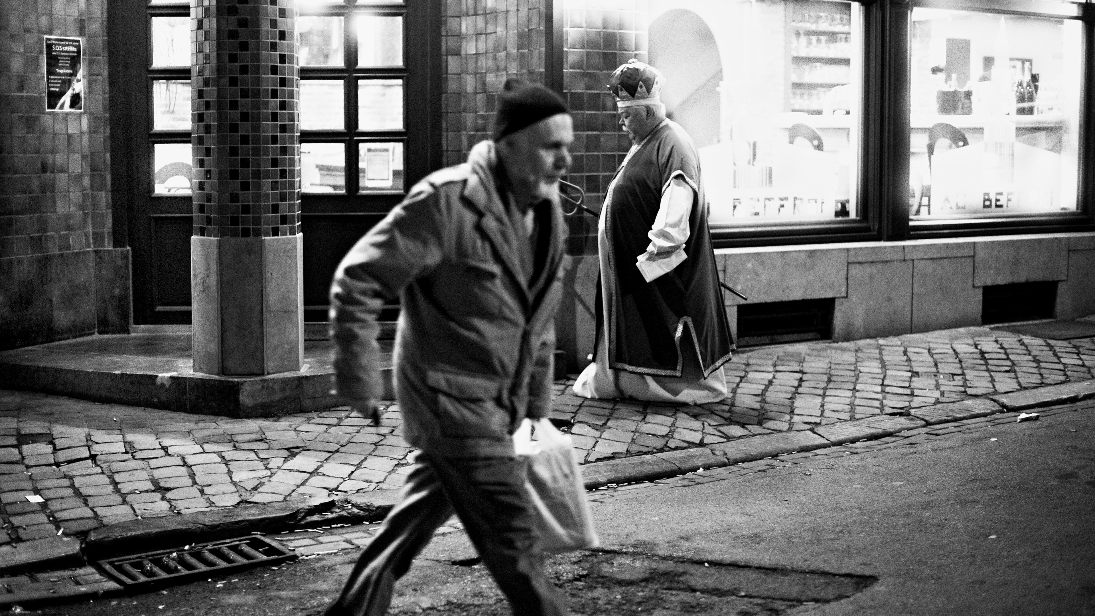 Black-and-white photo of a man walking past another dressed as a bishop on a cobblestone street at night.