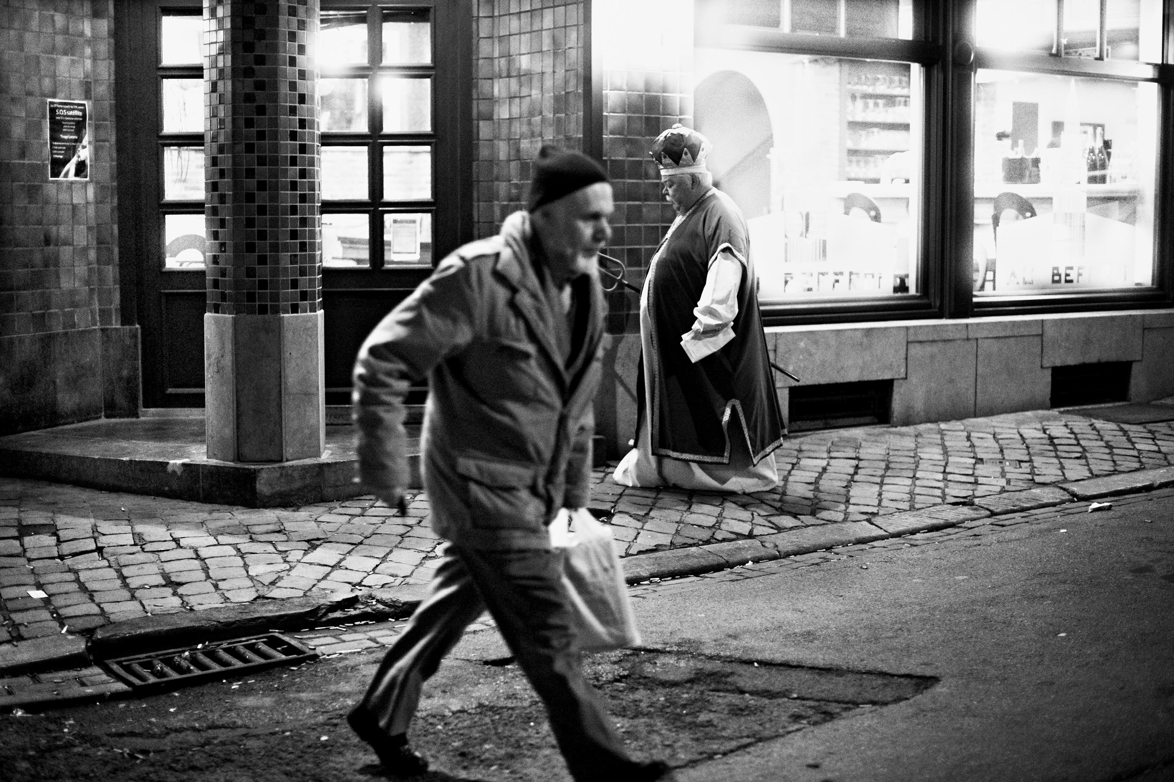 Black-and-white photo of a man walking past another dressed as a bishop on a cobblestone street at night.