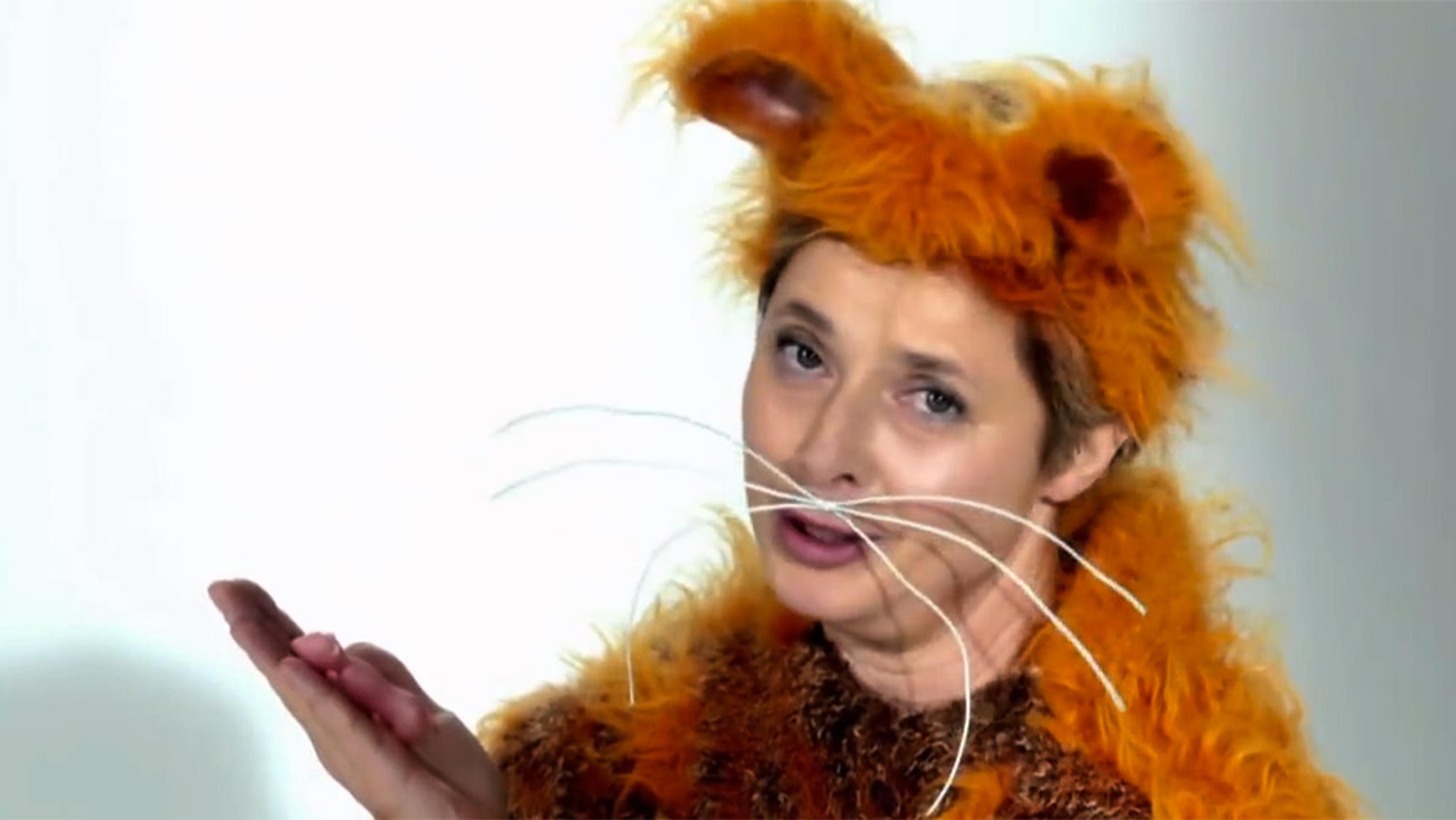 The actress Isabella Rossellini in an orange furry cat costume with whiskers, looking at the camera while gesturing with one hand against a plain background.