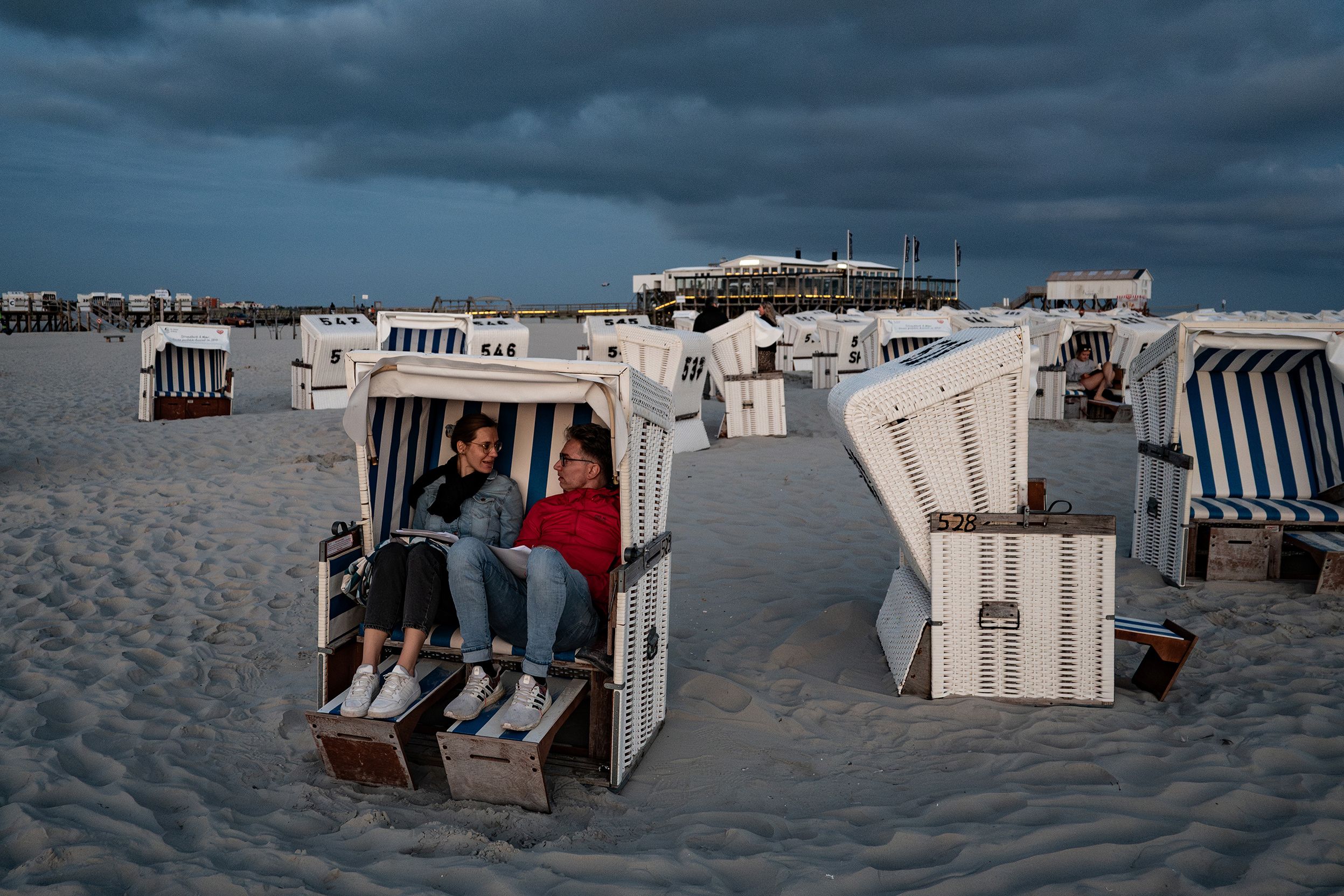 A couple sitting in a beach chair on a sandy shore during a cloudy evening surrounded by empty striped chairs.