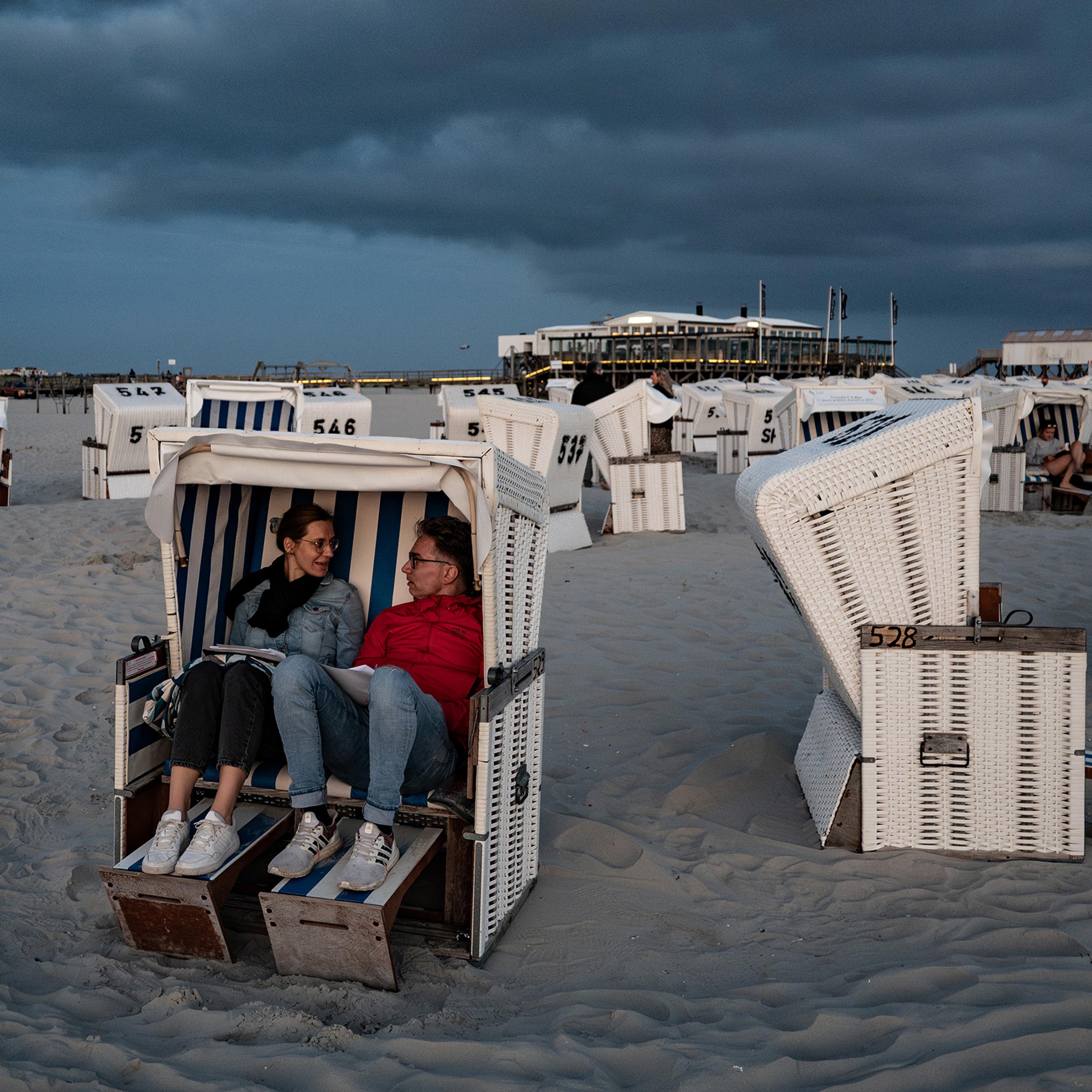 A couple sitting in a beach chair on a sandy shore during a cloudy evening surrounded by empty striped chairs.
