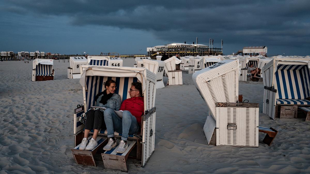 A couple sitting in a beach chair on a sandy shore during a cloudy evening surrounded by empty striped chairs.