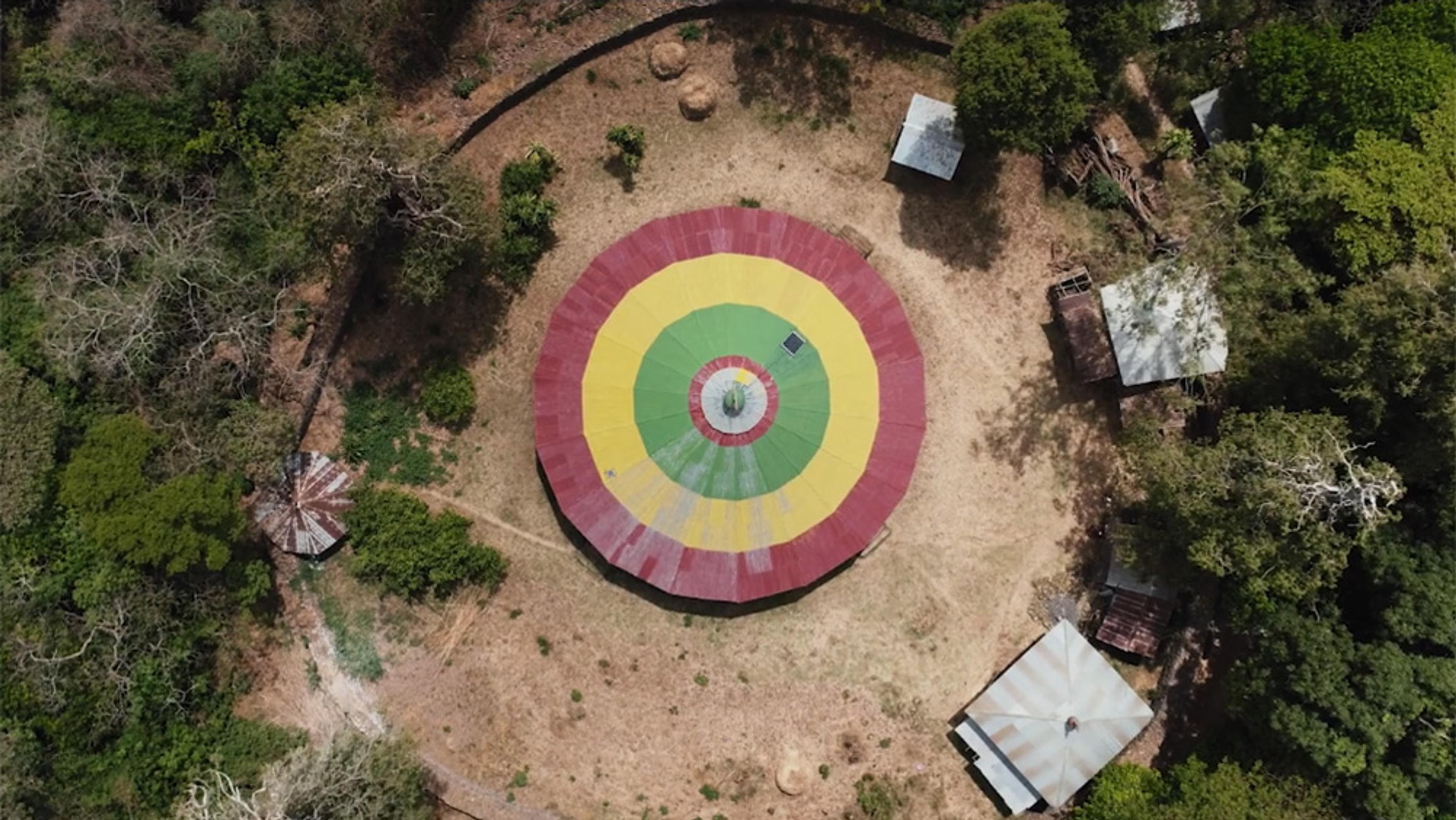 Aerial photo of a circular building with a red, yellow, and green roof surrounded by trees and smaller buildings in a clearing.