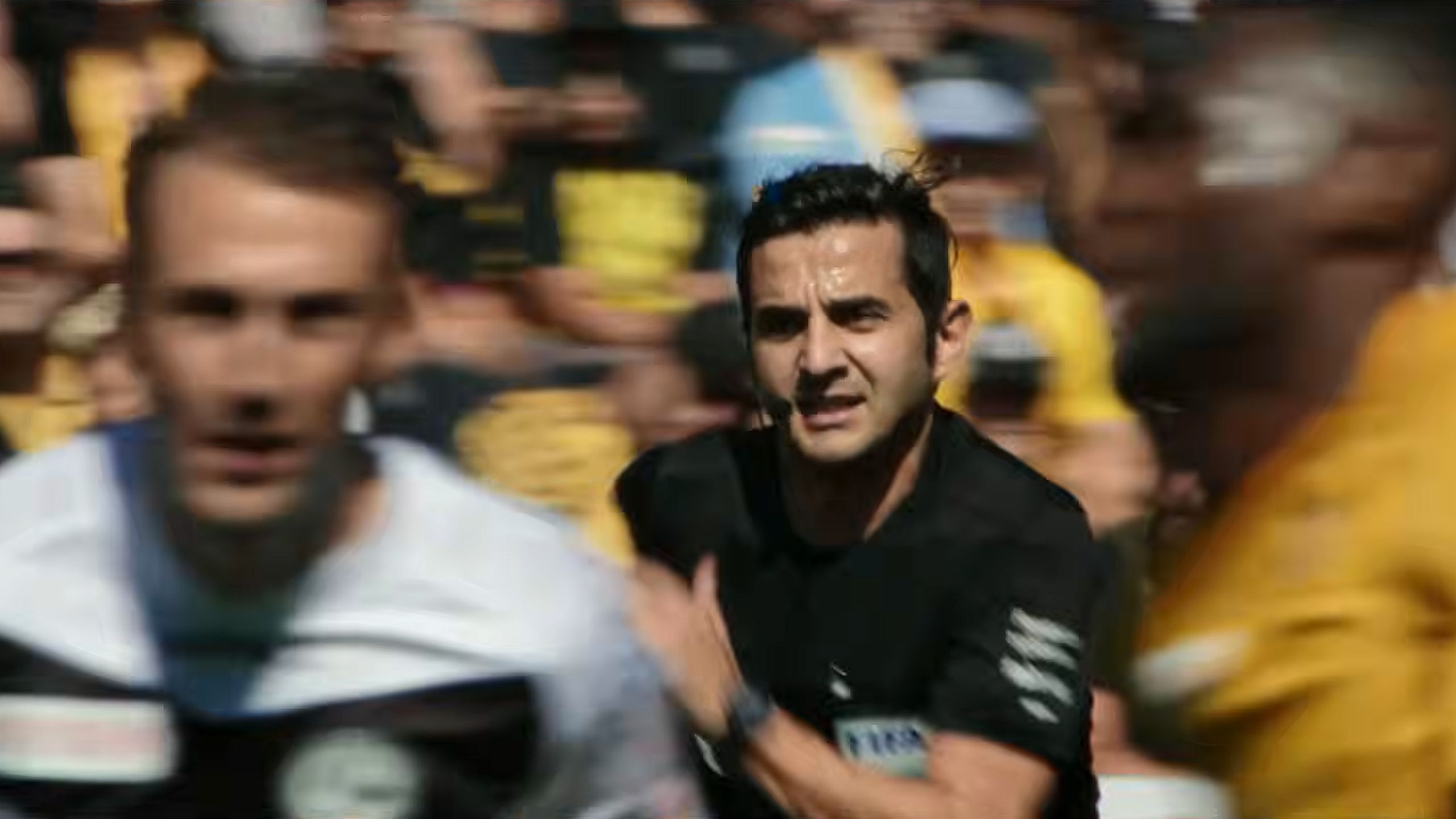 Photo of a football referee running on a pitch with blurred players and crowd in the background.