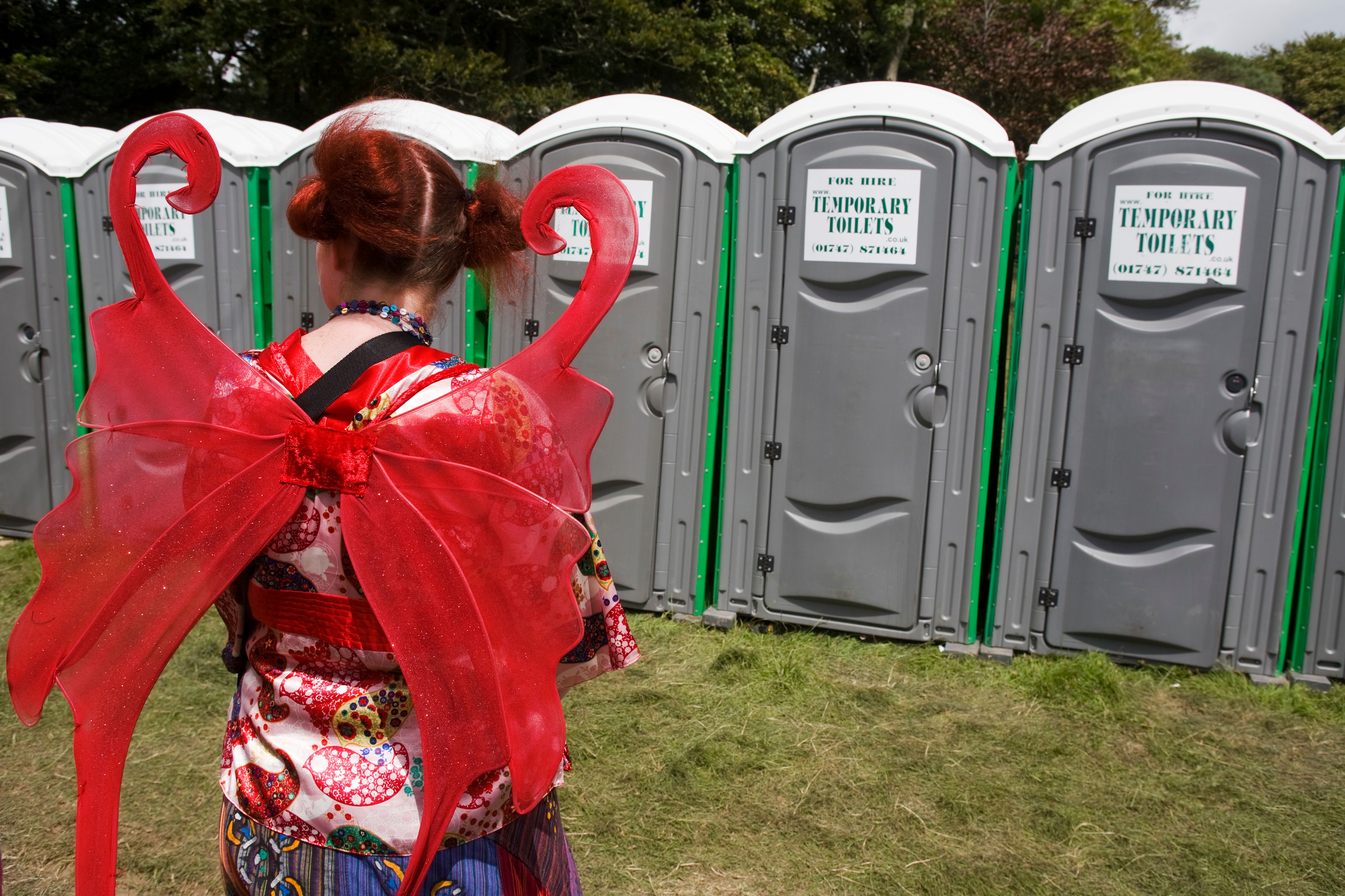 Photo of person in red fairy wings facing portable toilets outdoors.