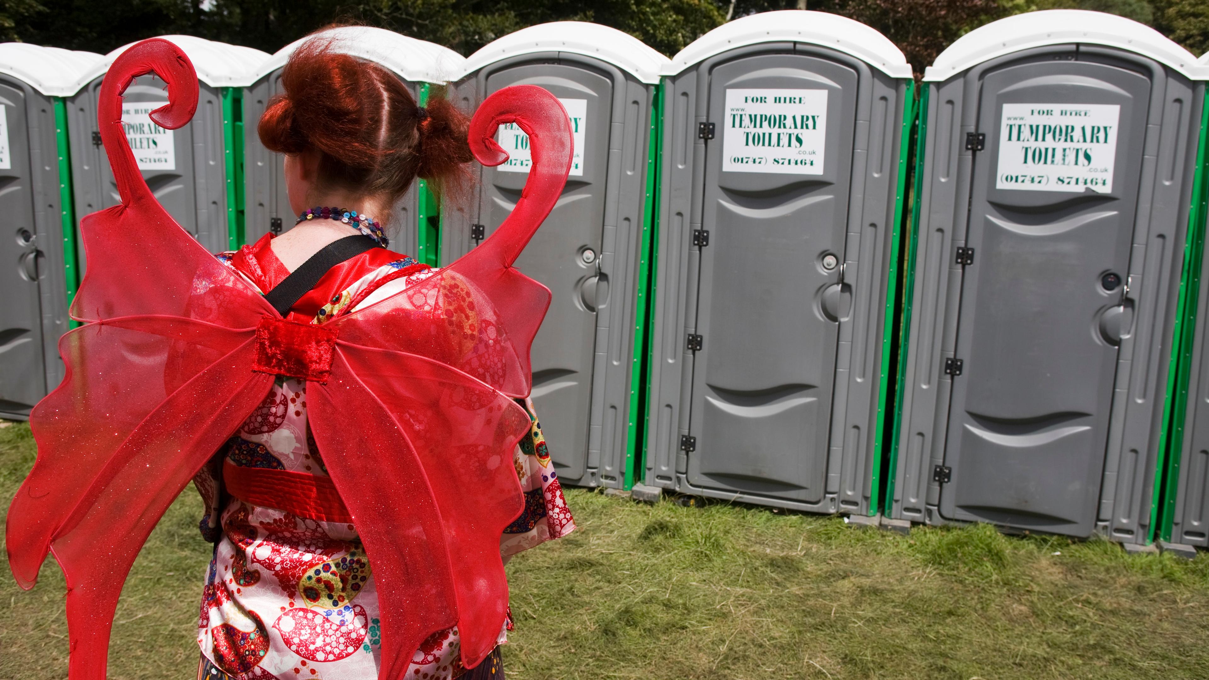 Photo of person in red fairy wings facing portable toilets outdoors.