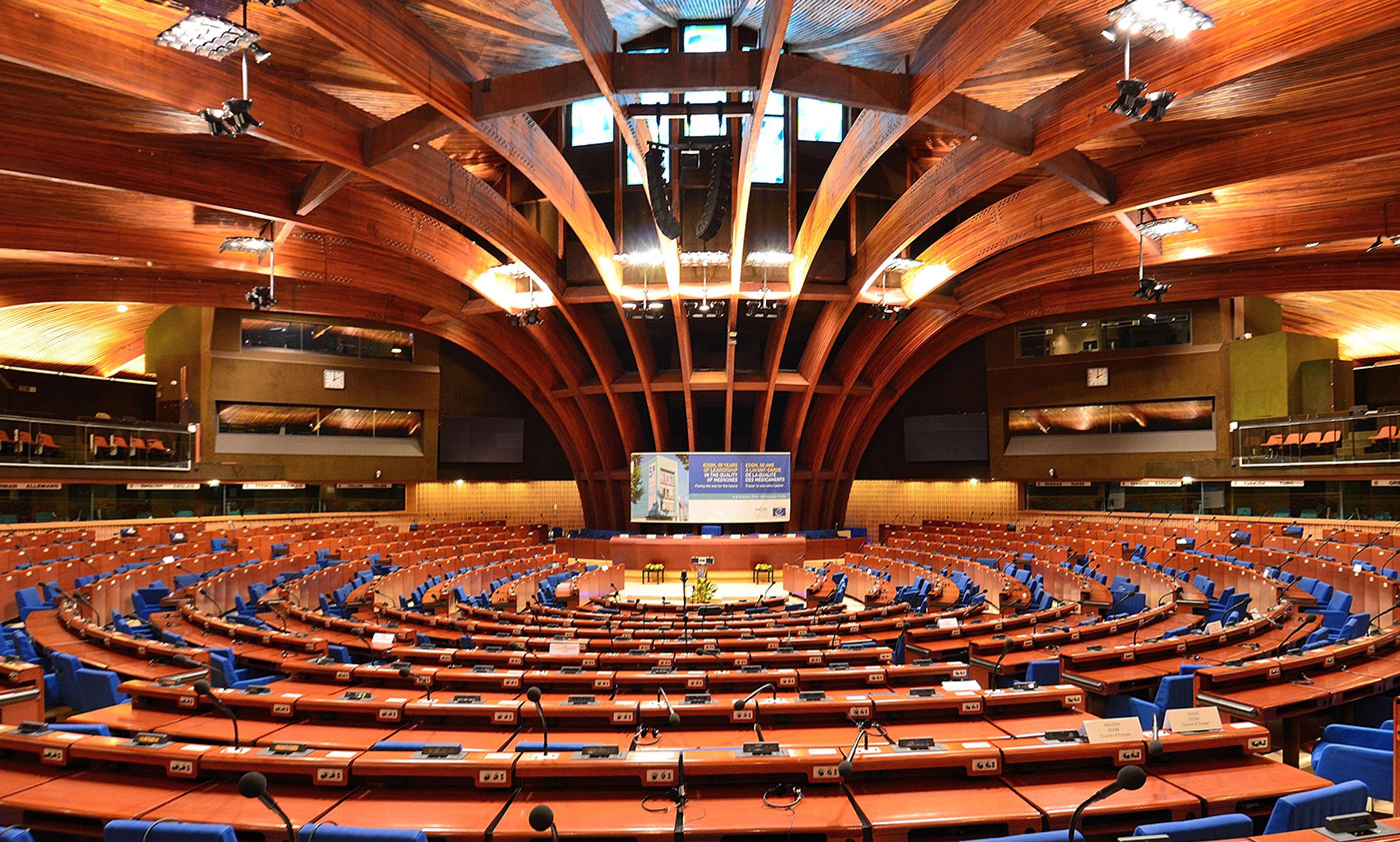 An empty, semi-circular parliamentary chamber with desks, blue chairs, and a unique wooden ceiling structure.