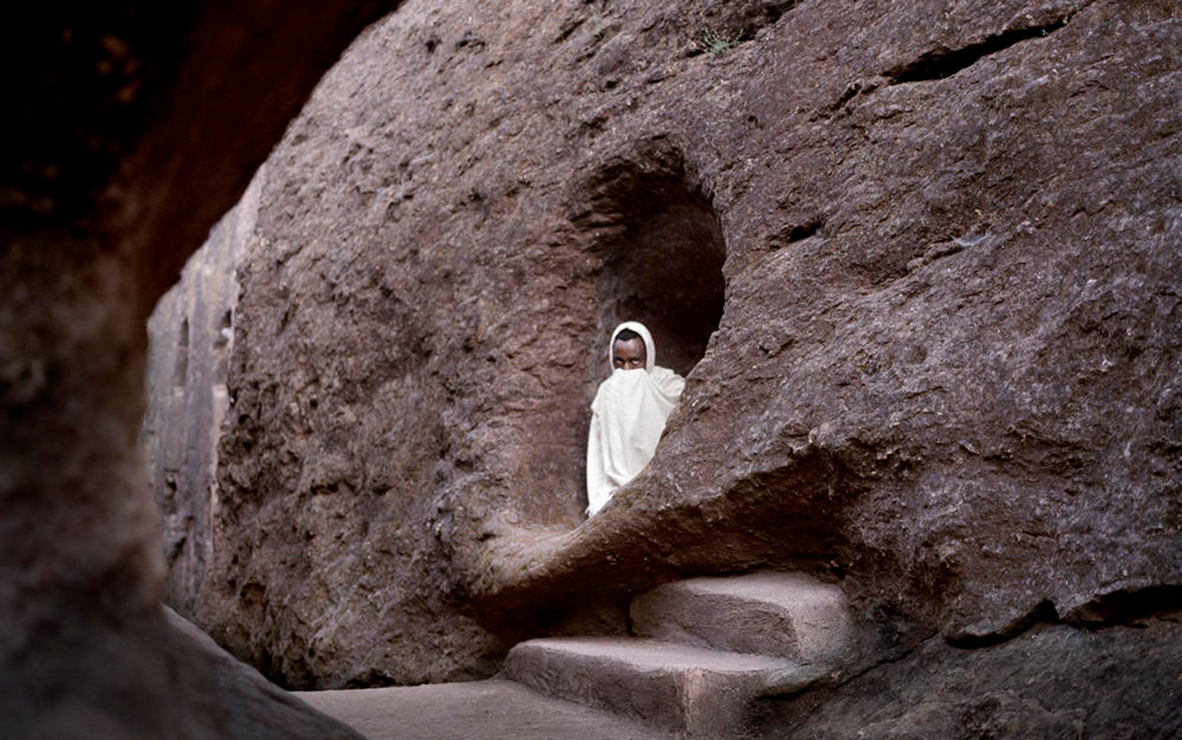A man dressed in white robes with his face obscured is seen in a cave entrance carved from rock