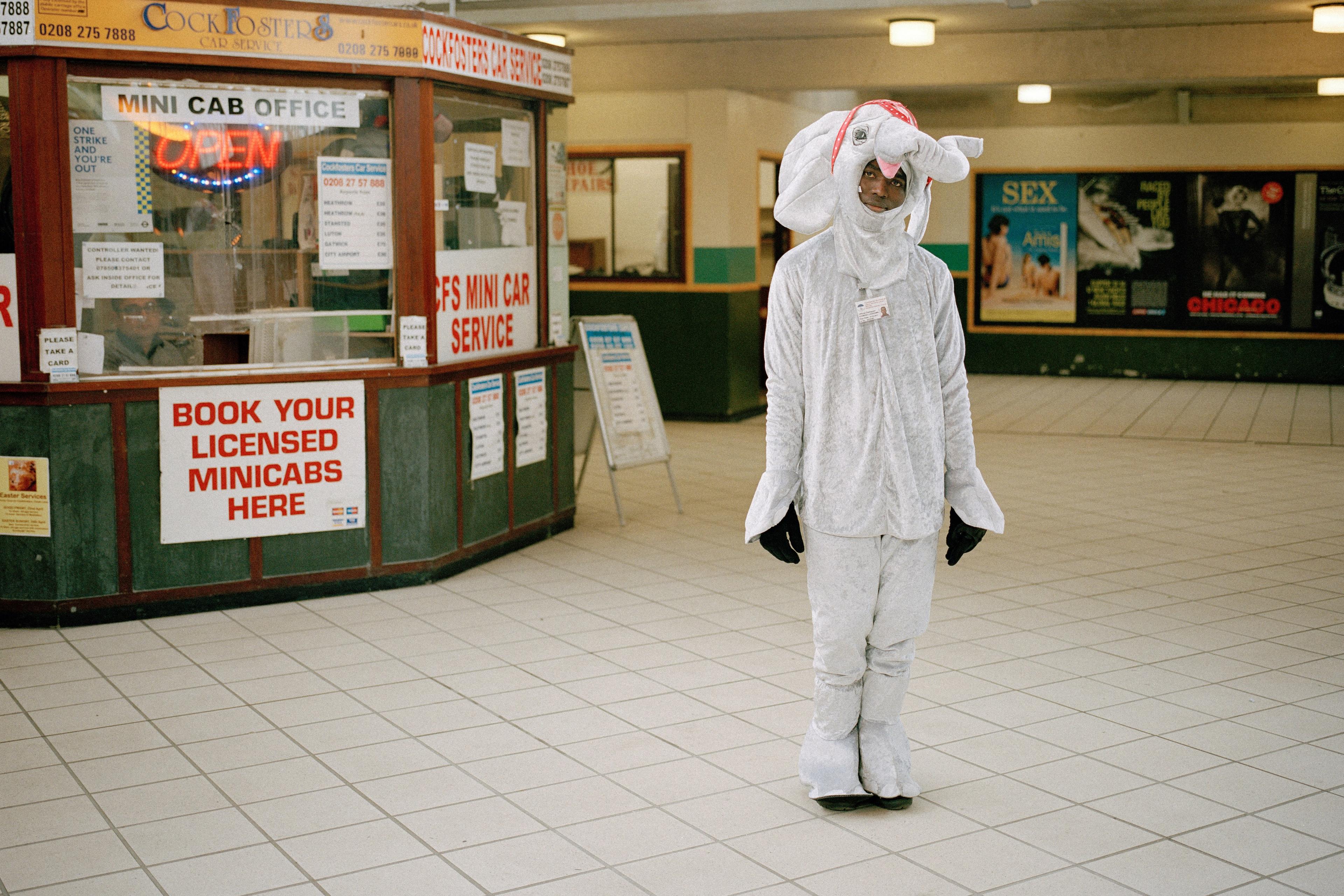 Photo of a person in an elephant costume standing in a tiled indoor area near a mini cab office with signs and posters.