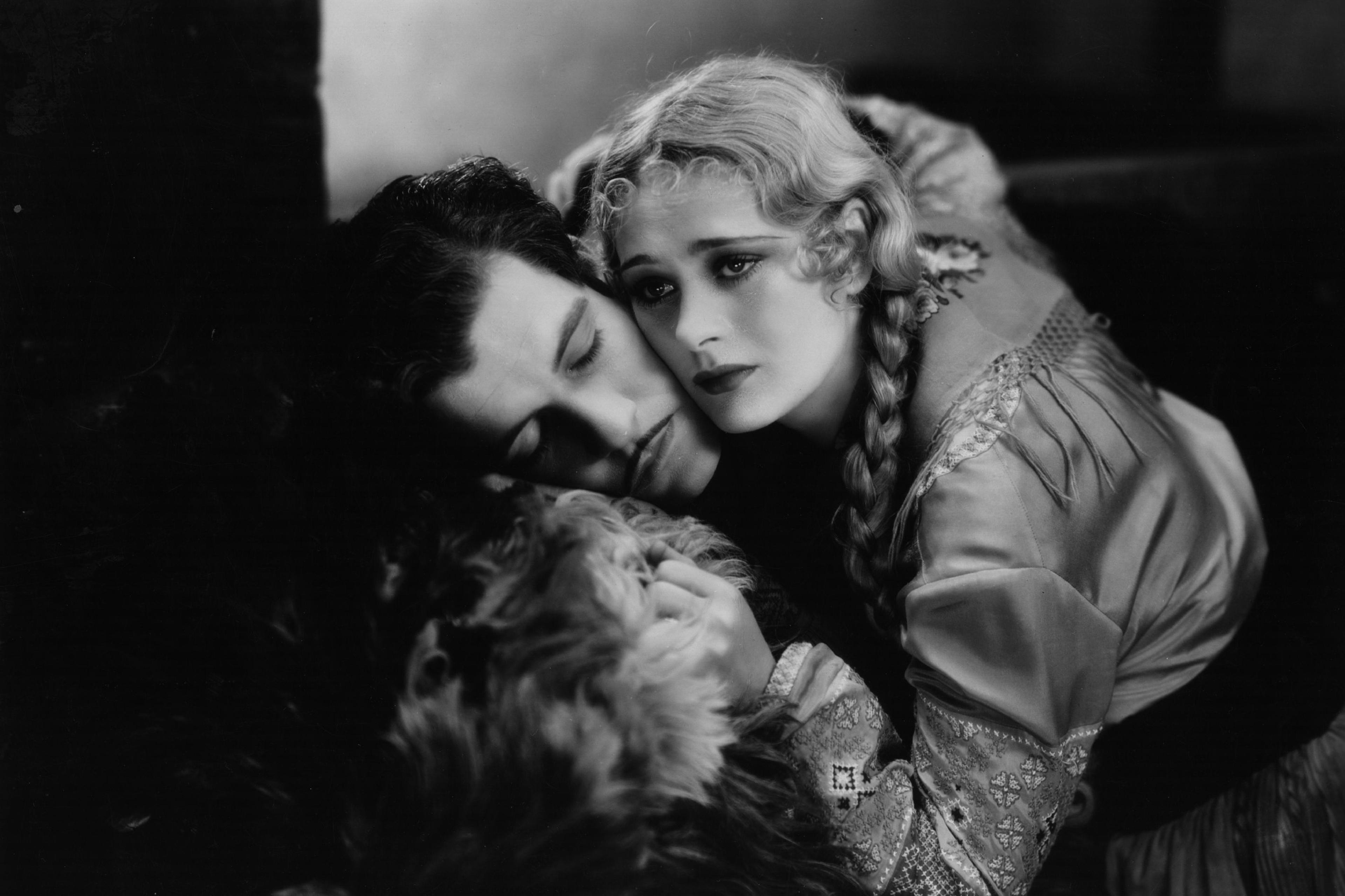 Black and white photo of a woman with braided hair cradling a man who is resting his head on her lap, both appear pensive.