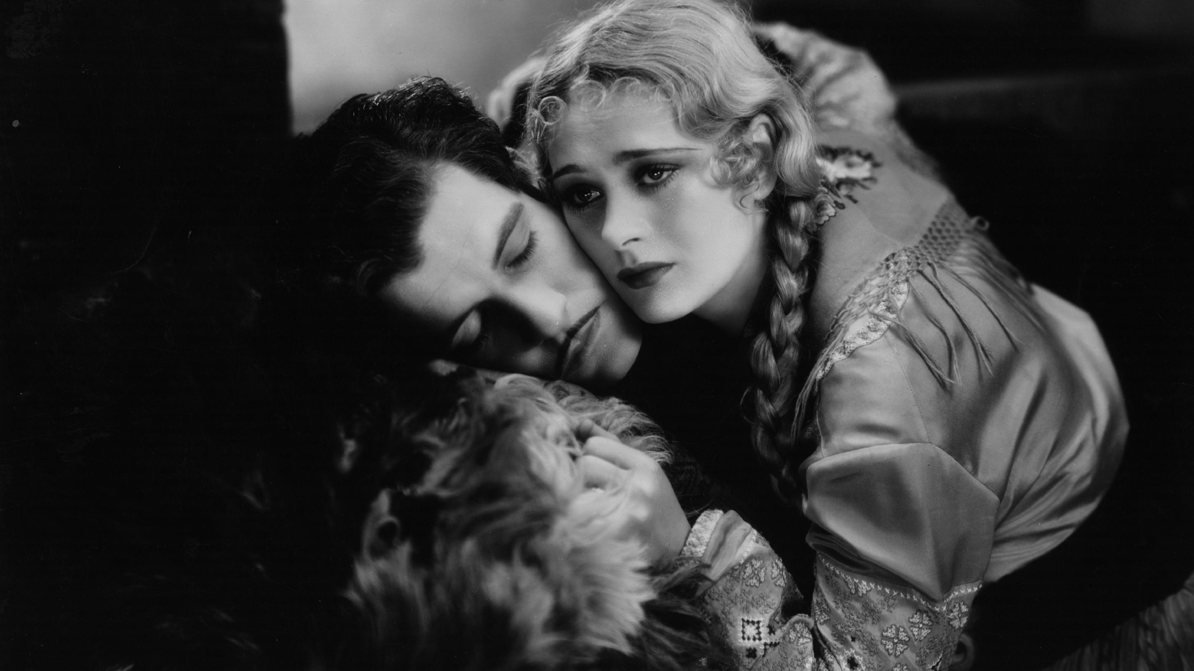 Black and white photo of a woman with braided hair cradling a man who is resting his head on her lap, both appear pensive.