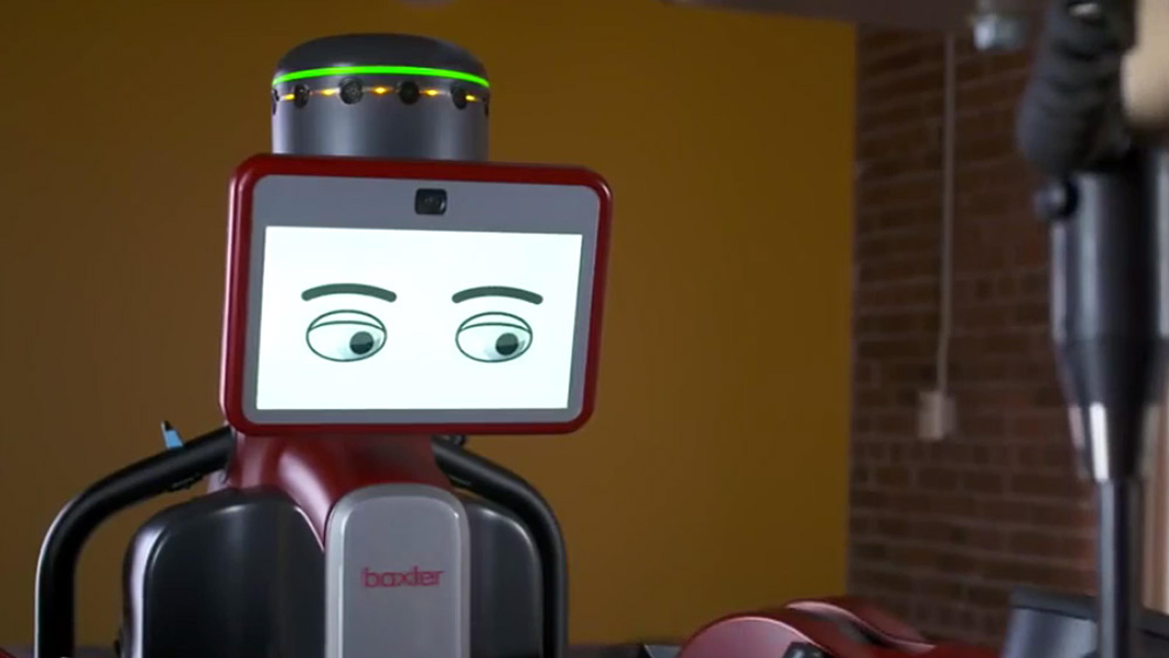A humanoid robot with a screen displaying cartoon eyes, set in a room with a yellow wall and dark brick wall.