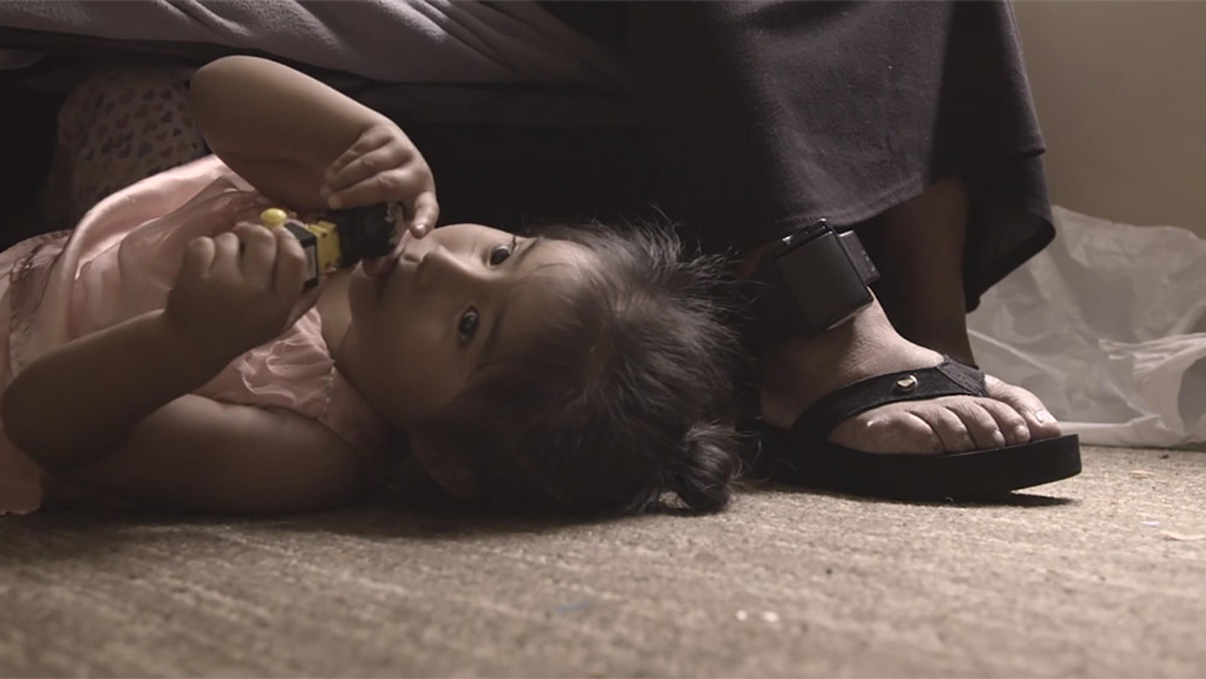 A young child lying on the floor playing, next to an adult wearing a black ankle monitor and sandal; seen from the knee down.