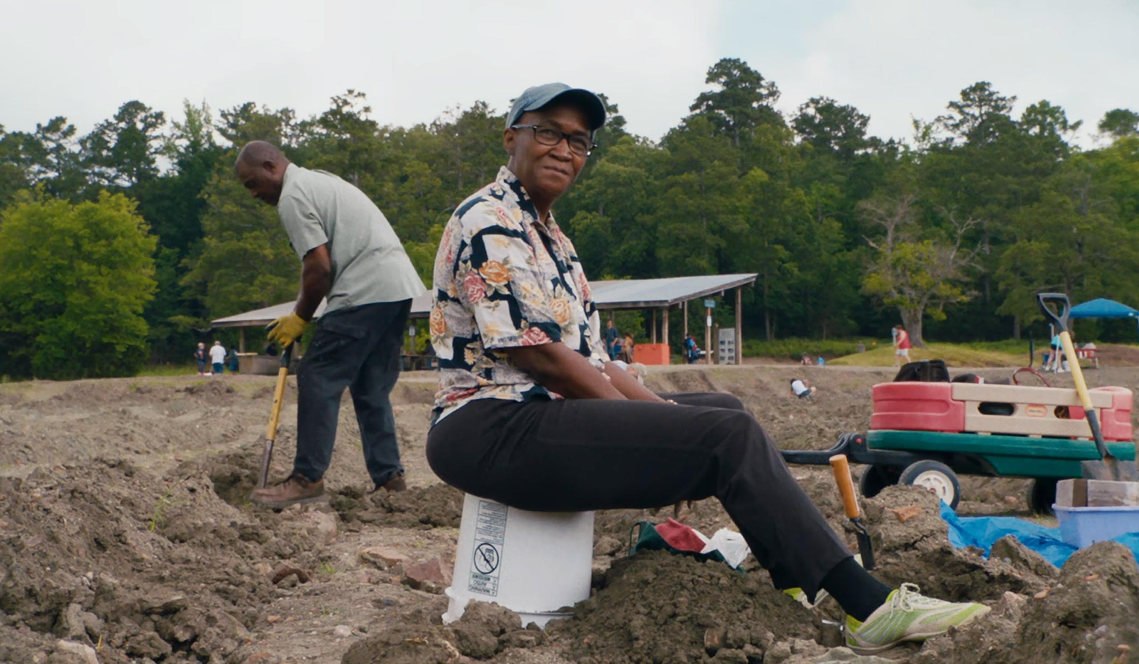 Person sitting on a bucket in a field, with another person digging nearby and trees in the background.