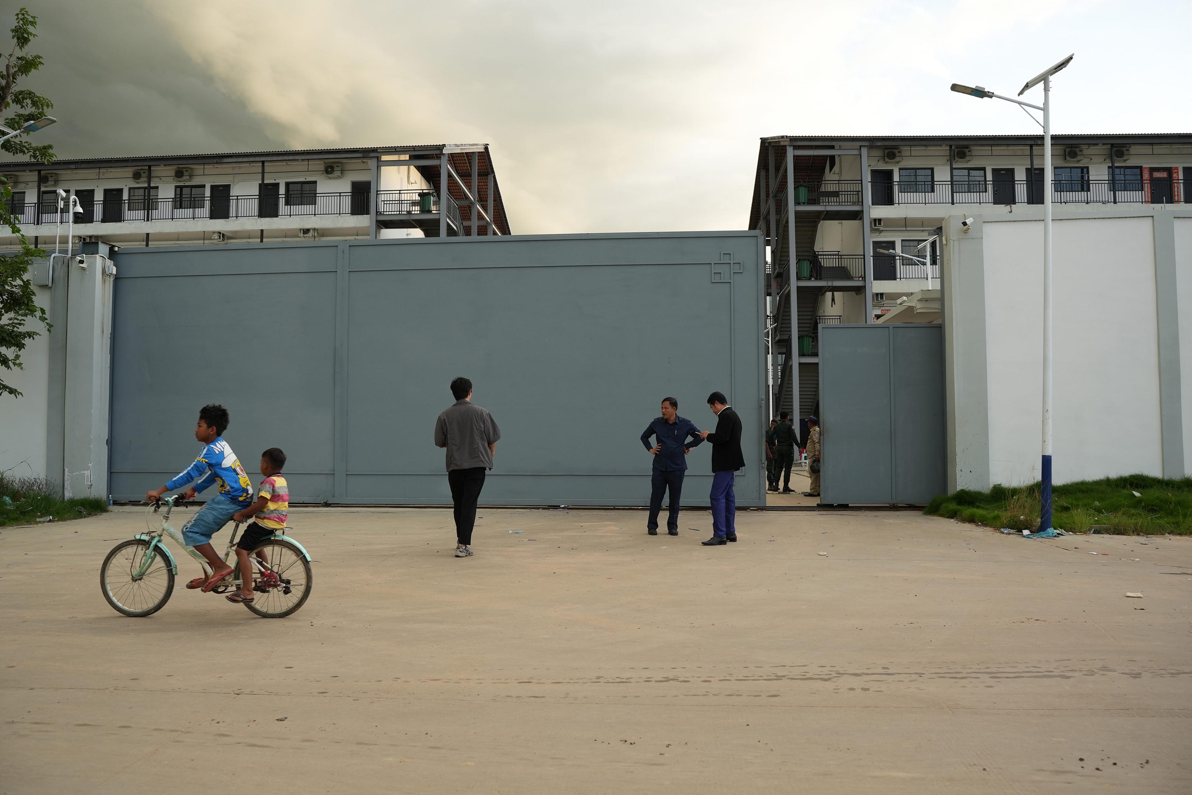 Two children on a bike passing a group of people standing outside a large metal gate with buildings in the background.